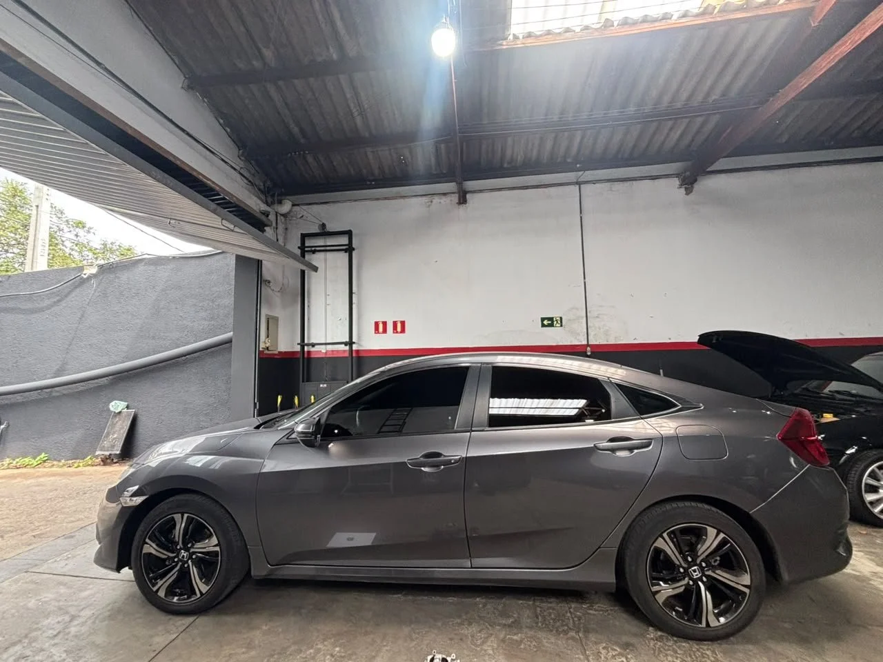 A gray Honda Civic parked inside a garage with an open trunk. The garage has a concrete floor and a white wall with red and black accents. Part of another car is visible on the right side.