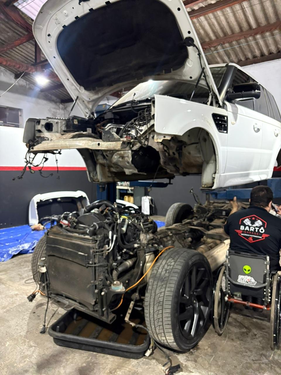 A white SUV is raised on a lift with its hood open, revealing the engine compartment, while a person in a wheelchair works on the disassembled engine in a garage or repair shop.