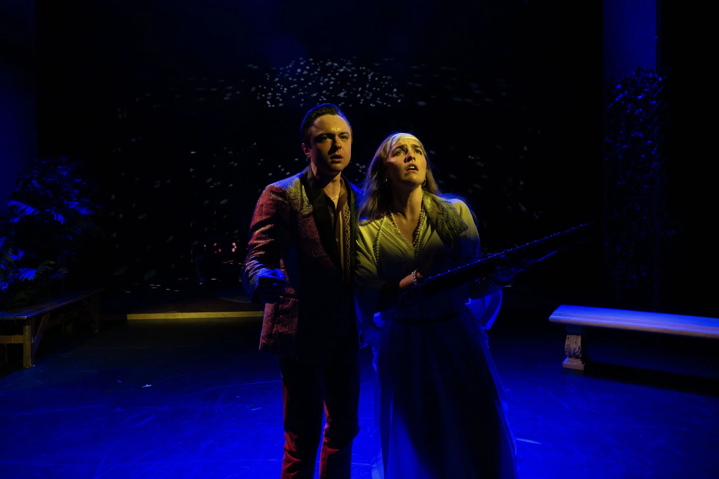 Two actors on stage, a man and a woman, who appear worried or distressed, illuminated by dramatic blue and yellow stage lighting, with a dark background and some stage props on either side.