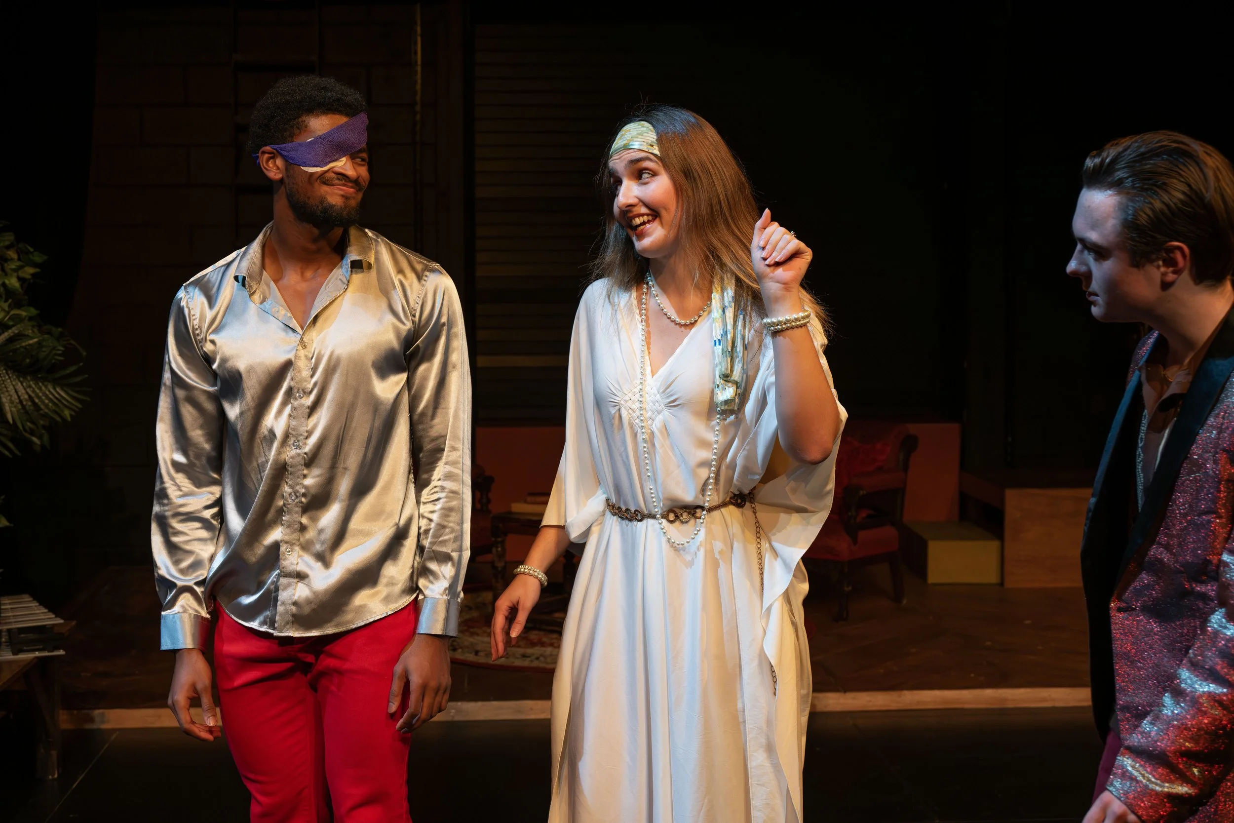 Three people on stage, one woman in the center wearing a white dress and headband, smiling and gesturing, with two men on either side, one blindfolded, smiling.
