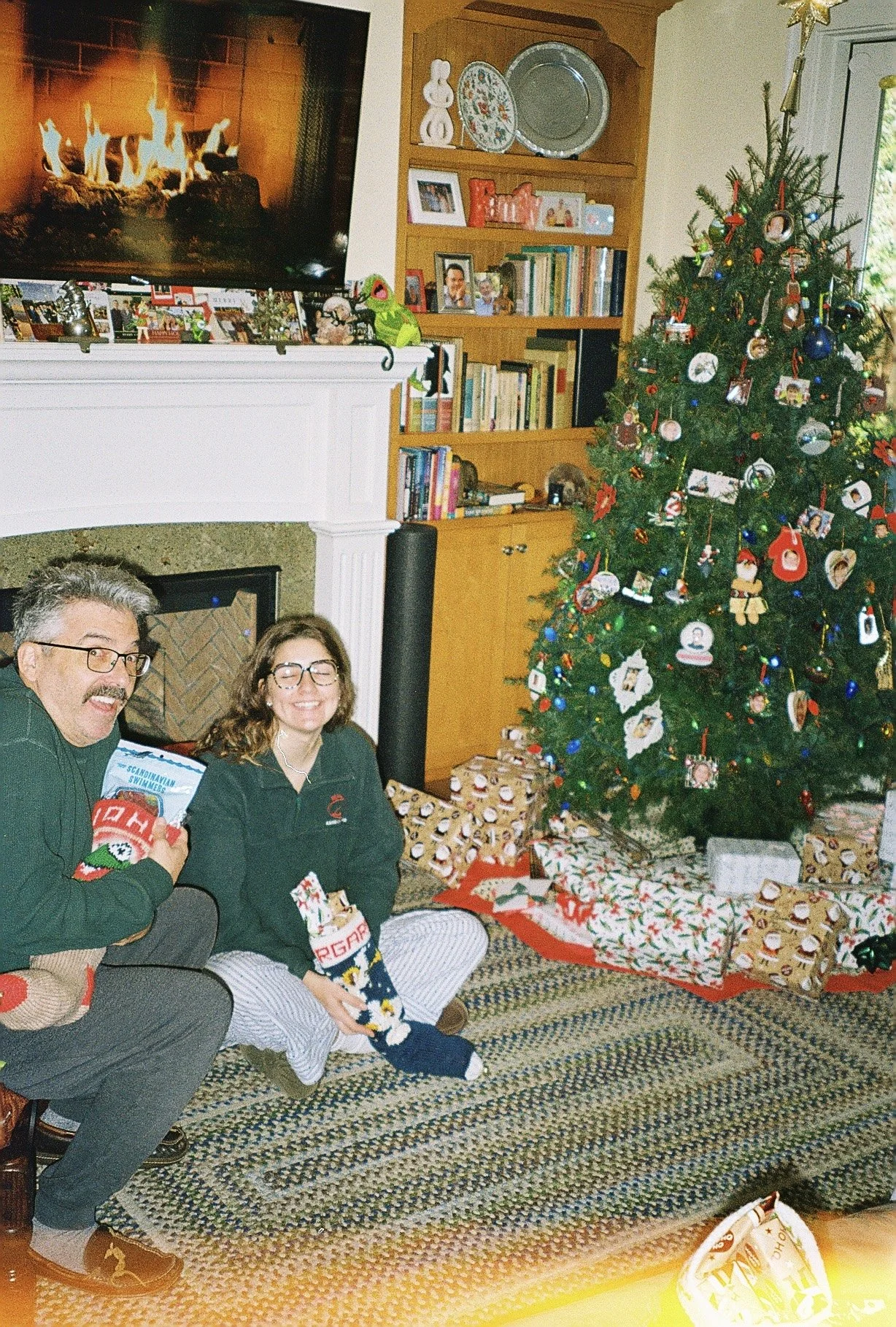 A living room decorated for Christmas with a large Christmas tree adorned with ornaments and lights, surrounded by wrapped presents. Two people are sitting on the carpet in front of the tree, smiling and holding gifts. A fireplace with a television m