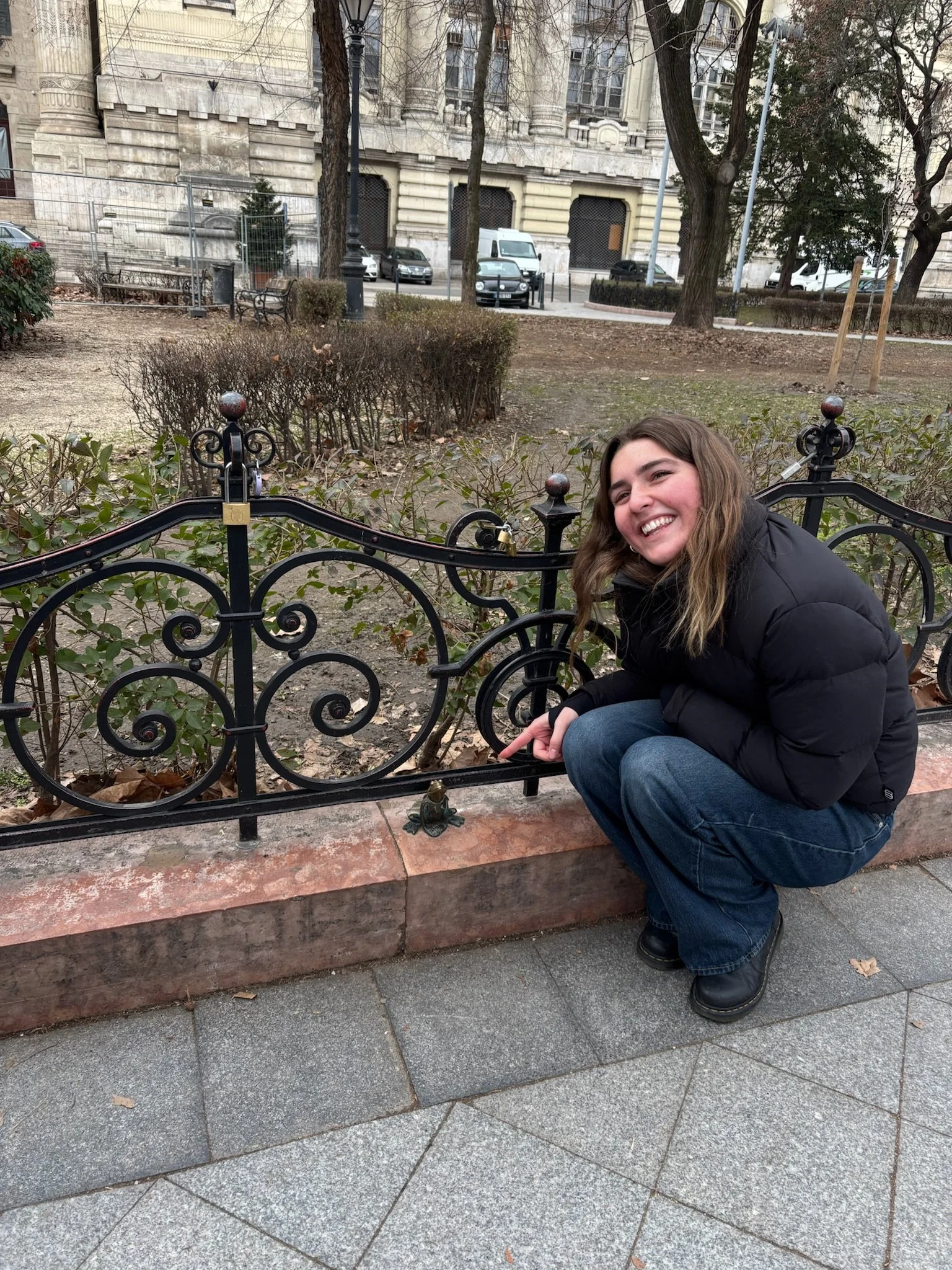 A smiling young woman with brown hair in a black jacket and blue jeans crouches on the sidewalk next to a decorative metal fence and a small frog statue at the base of the fence in an urban park setting with trees, bushes, and parked cars in the background.