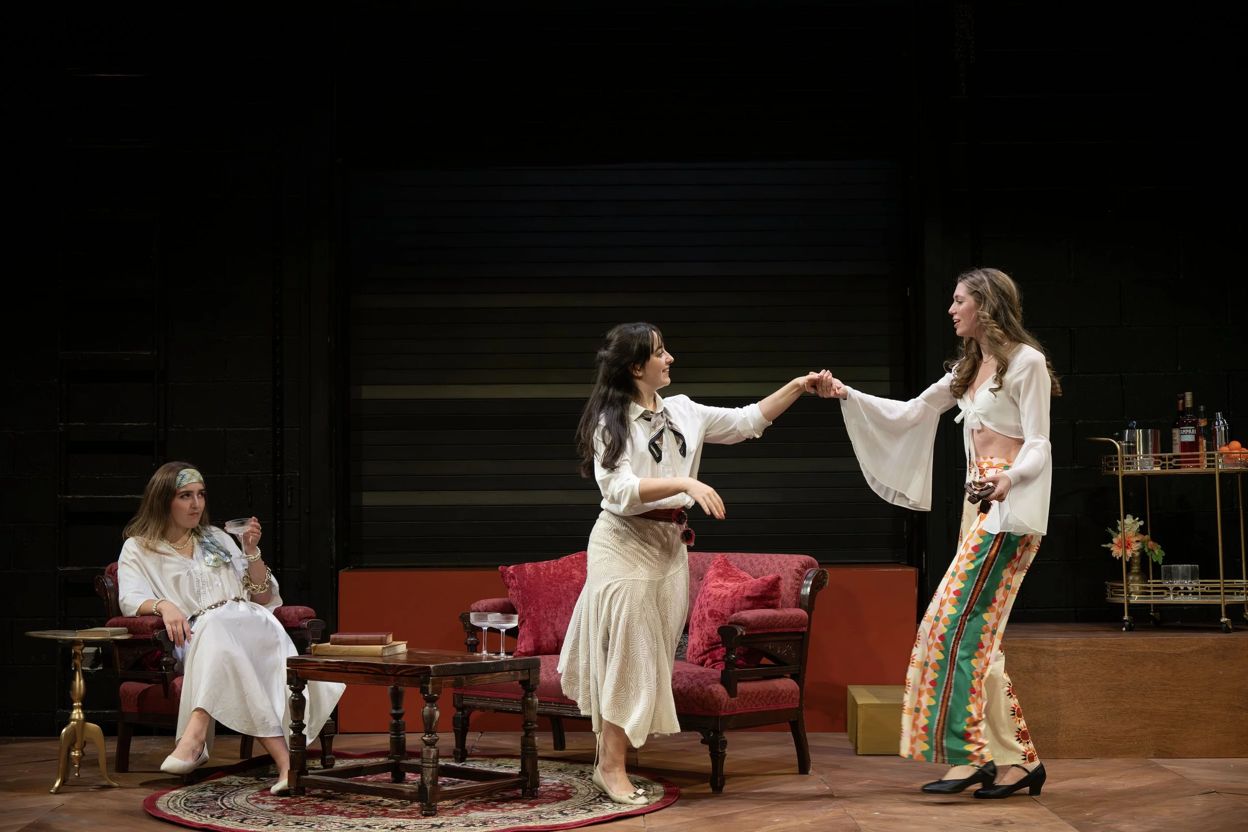 Four women on stage in a theatrical production, two are standing and holding hands, while two are seated carefully observing the scene. The set is styled like a living room with vintage furniture, books, and decorative items.