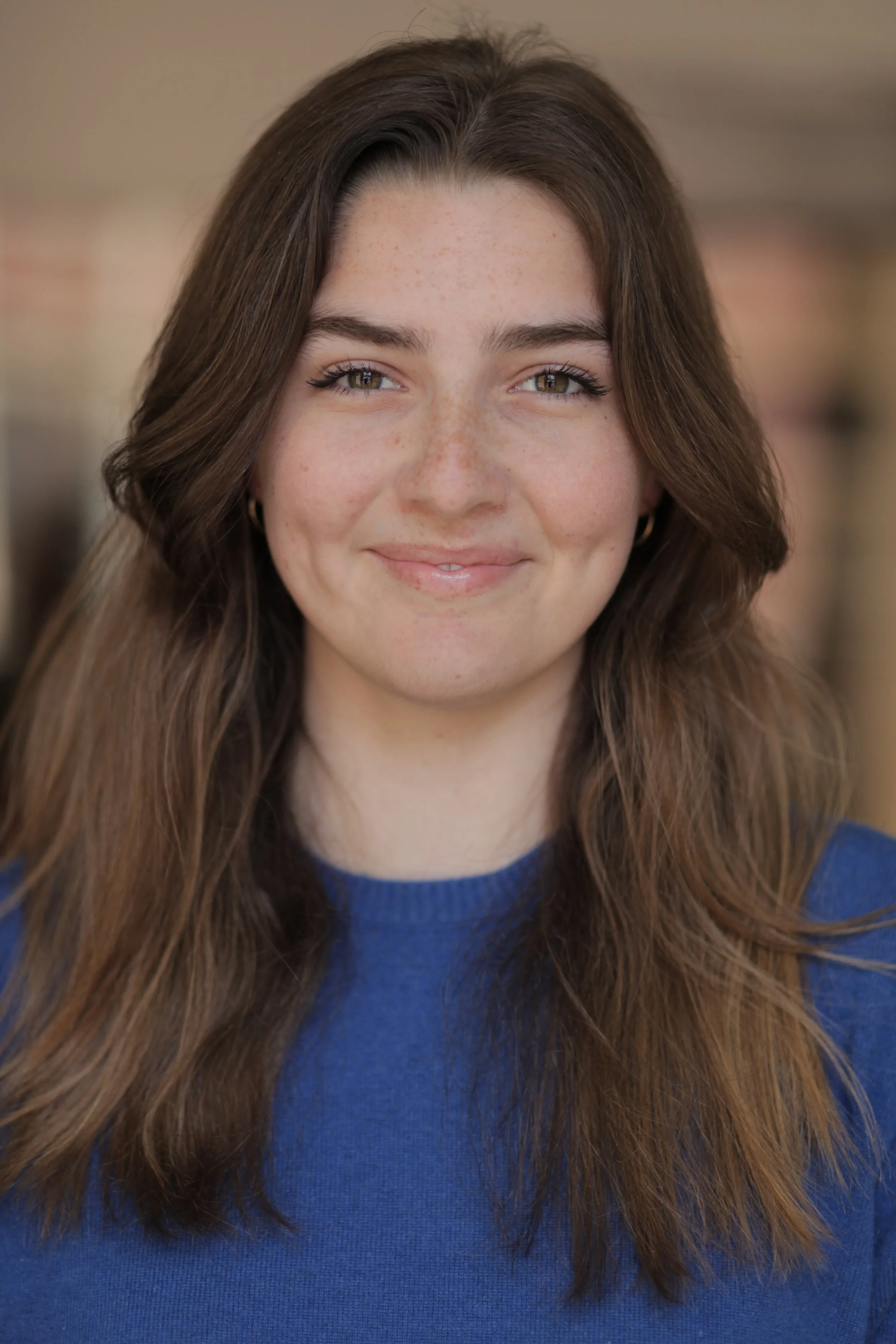 Close-up of a young woman with long brown hair, freckles, and brown eyes smiling at the camera, wearing a blue sweater.
