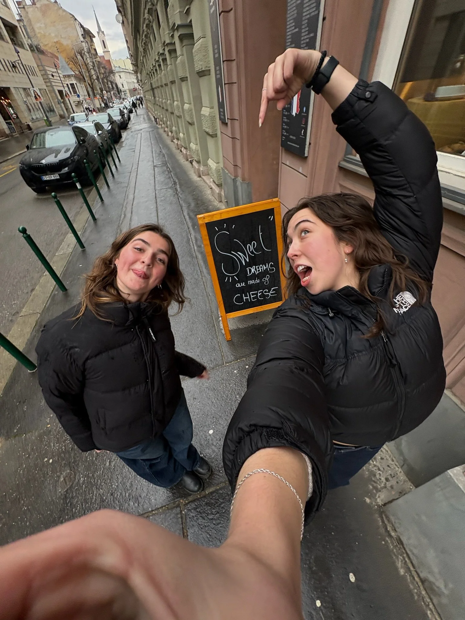 Two young women taking a selfie on a city sidewalk, with a sign behind them that reads "Sweet Dreams are made of Cheese." One woman is pointing up at her thumb, and the other woman is sticking her tongue out.