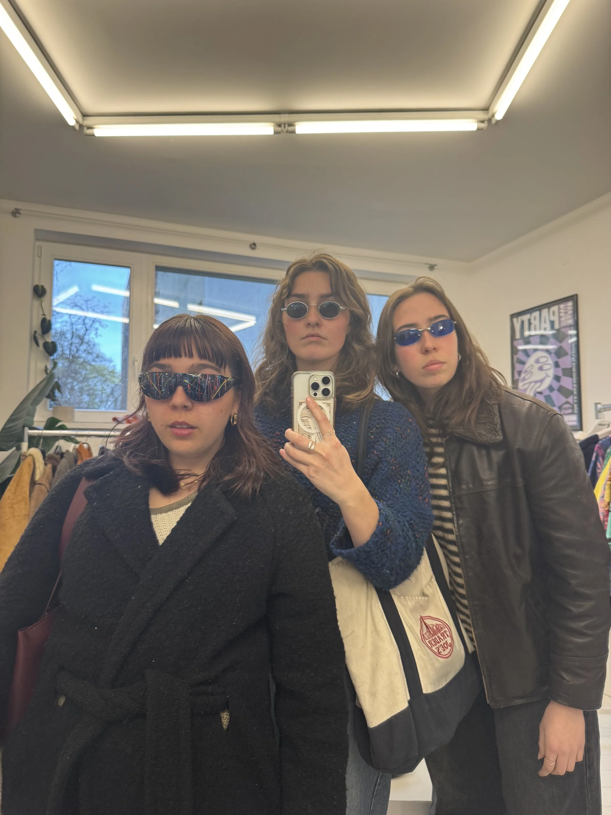 Three young women taking a mirror selfie in a room with a window and clothing racks. They are all wearing sunglasses and have different hairstyles.