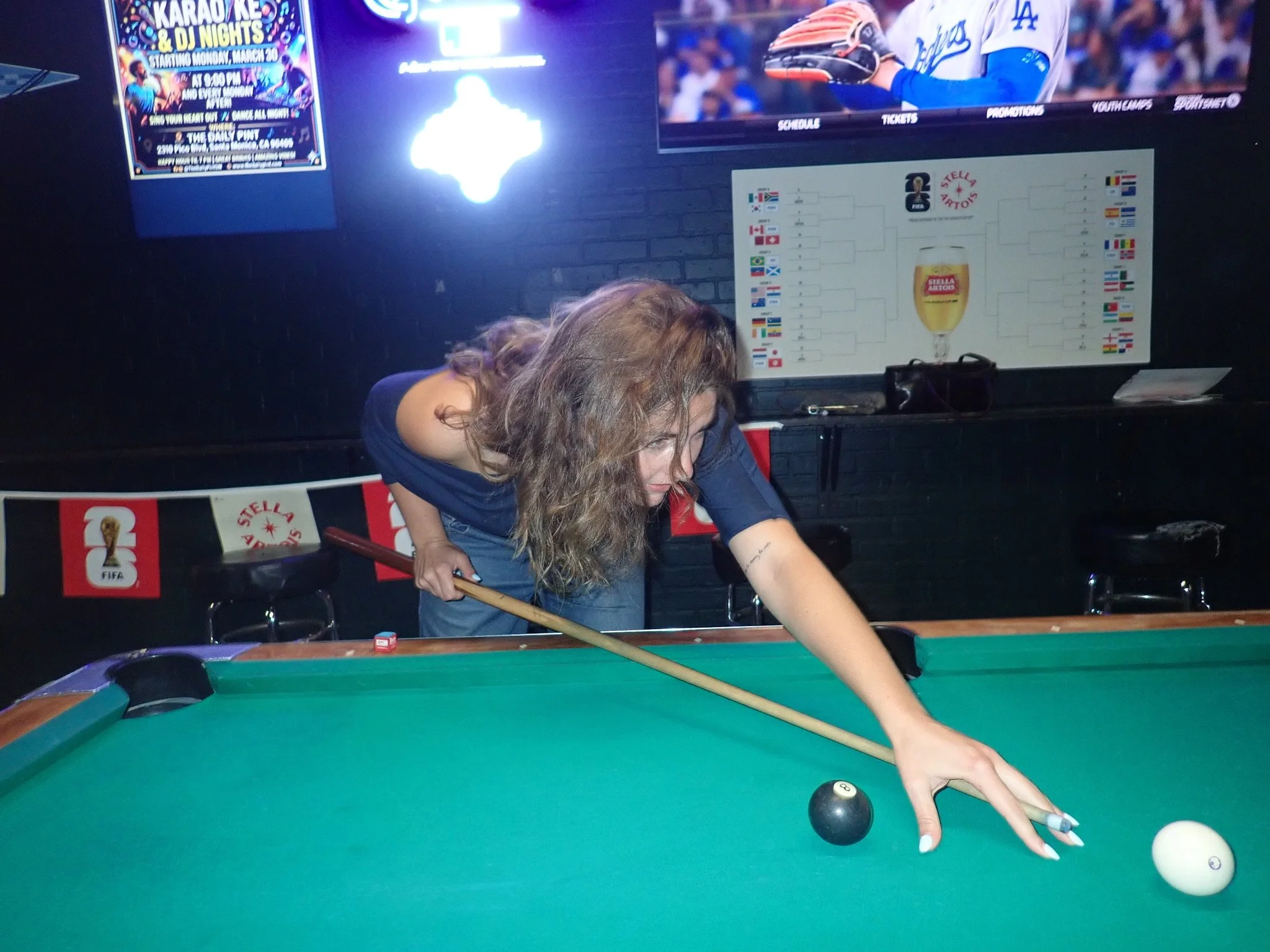 A woman with curly hair playing pool at a bar or pool hall, with multiple TV screens and a tournament bracket on the wall behind her.