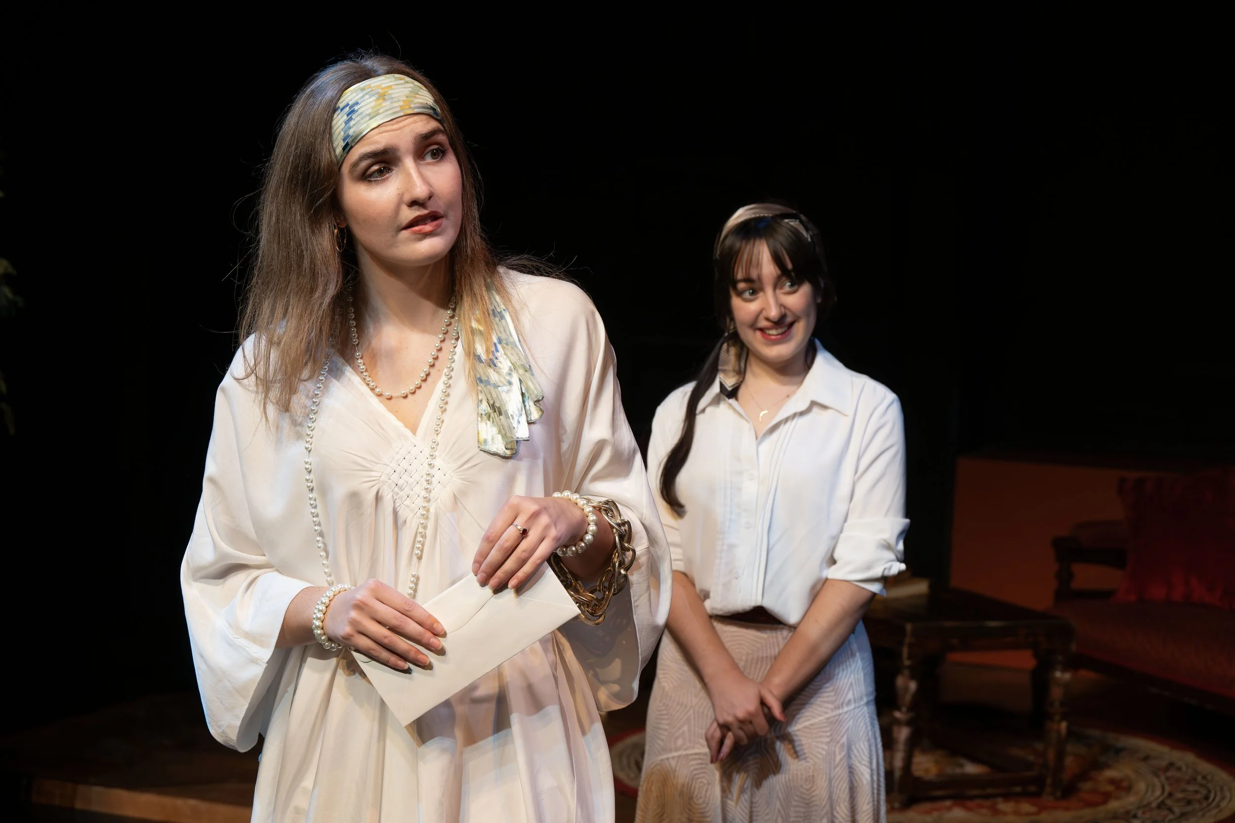 Two women standing indoors under stage lighting, one in the foreground wearing a headscarf, pearl necklace, and cream-colored dress holding a paper, the other smiling woman in the background wearing a white blouse and striped skirt.