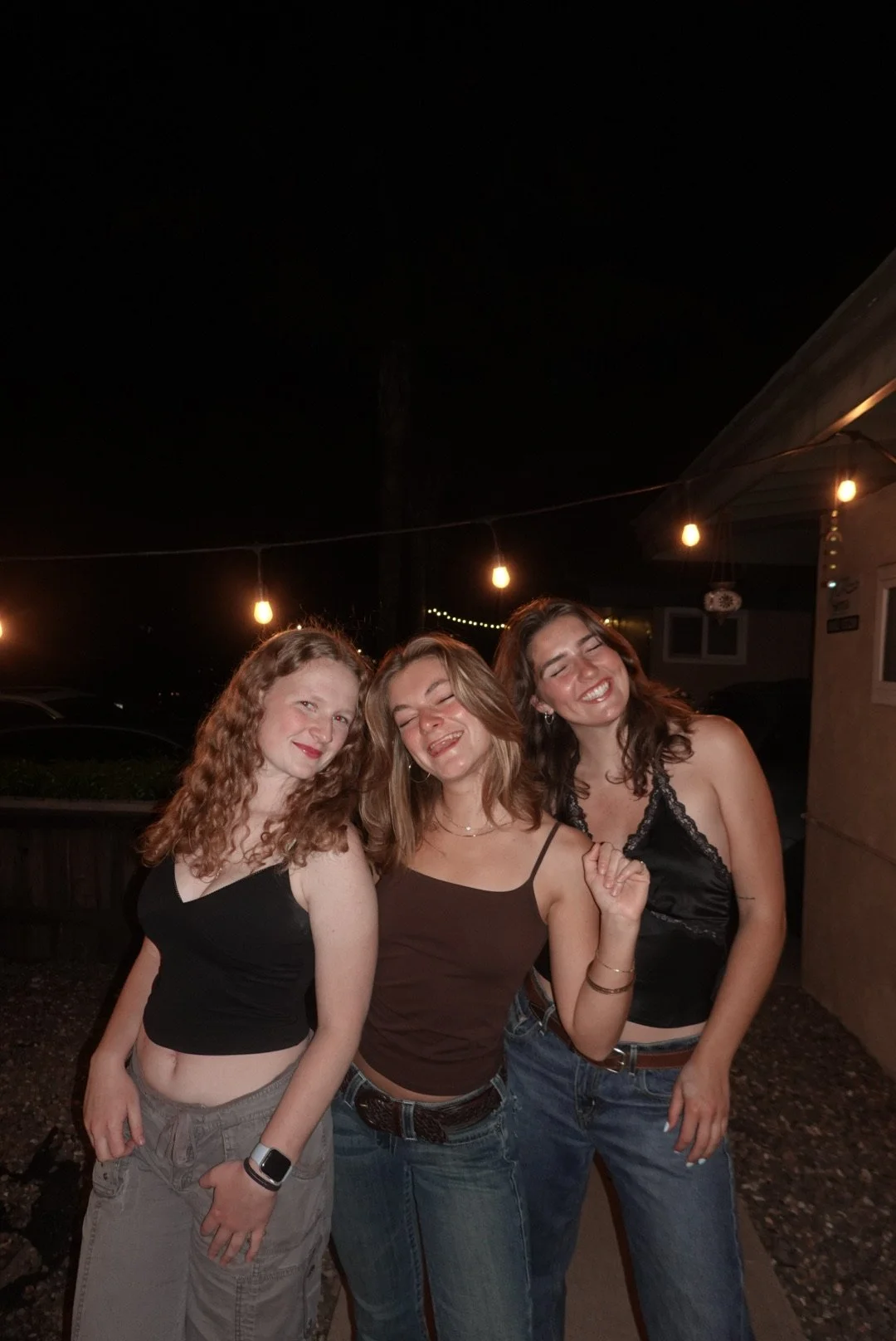 Three young women celebrating at night, smiling and posing outdoors under string lights