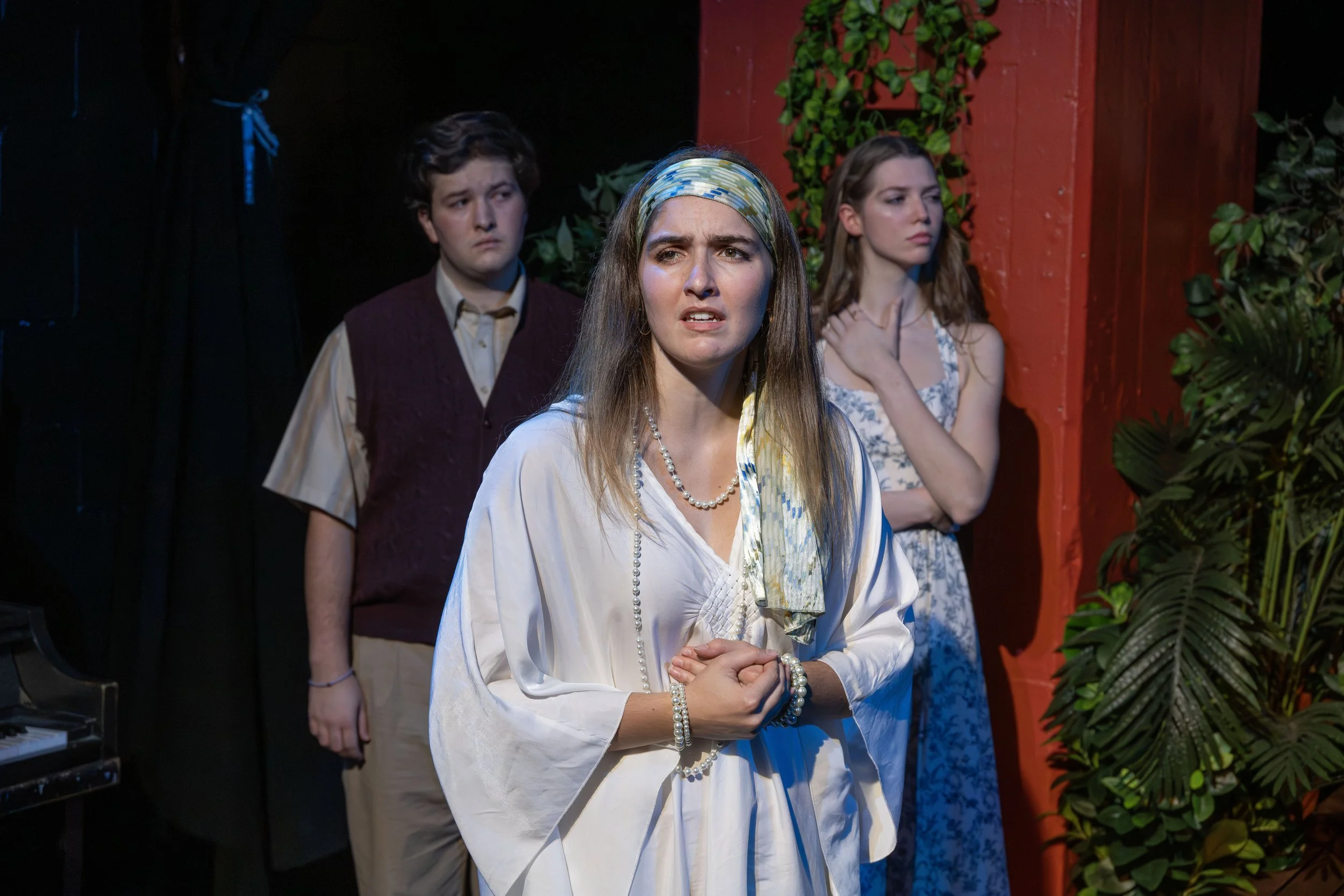 Three young women standing on a stage, with one in front looking concerned or confused, and two others in the background. The foreground woman wears a light-colored dress, pearl necklace, and headscarf. The background women wear vintage-style dresses