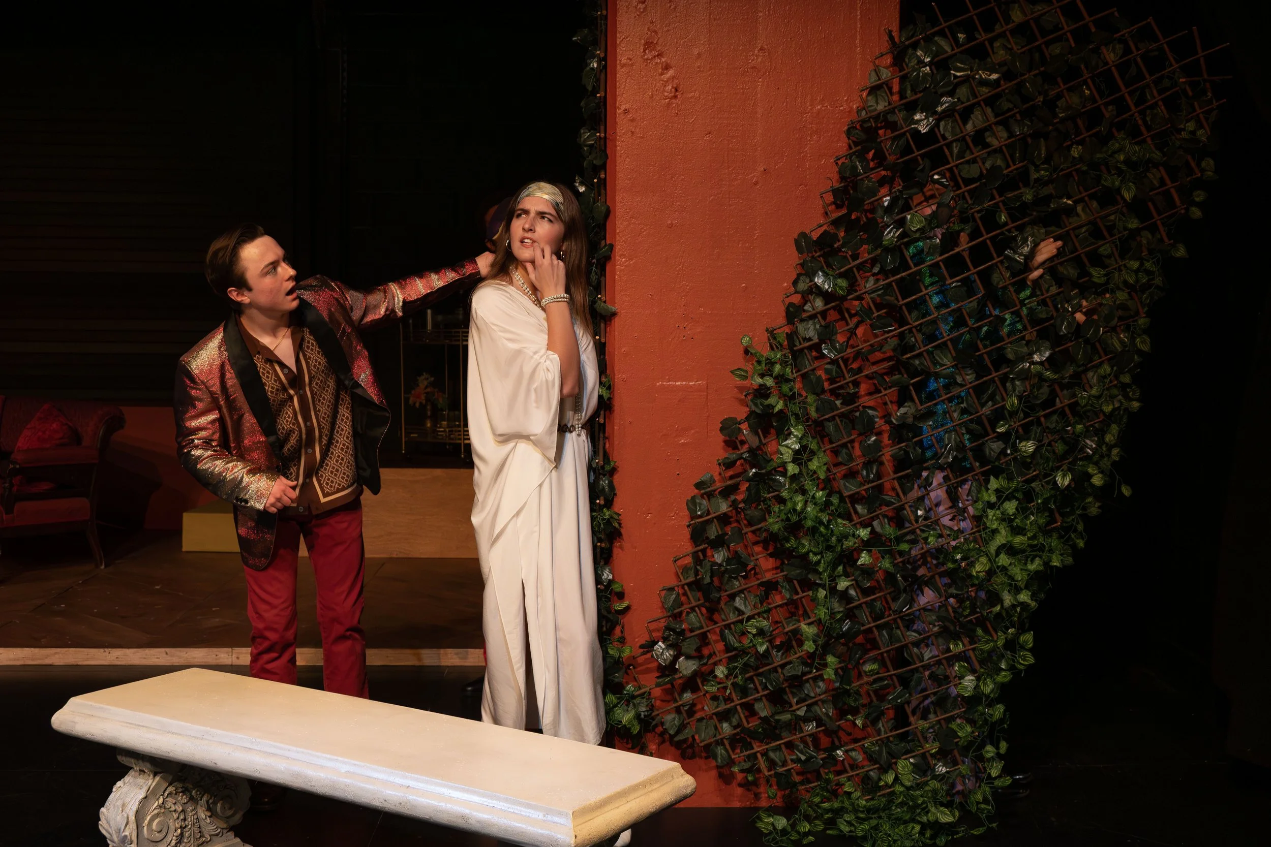 A young man and woman are on stage, the man is wearing a shiny, patterned blazer and red pants, and the woman is dressed in a white gown with pearls. The man has his arm around the woman's shoulders, and they both appear to be in a theatrical perform