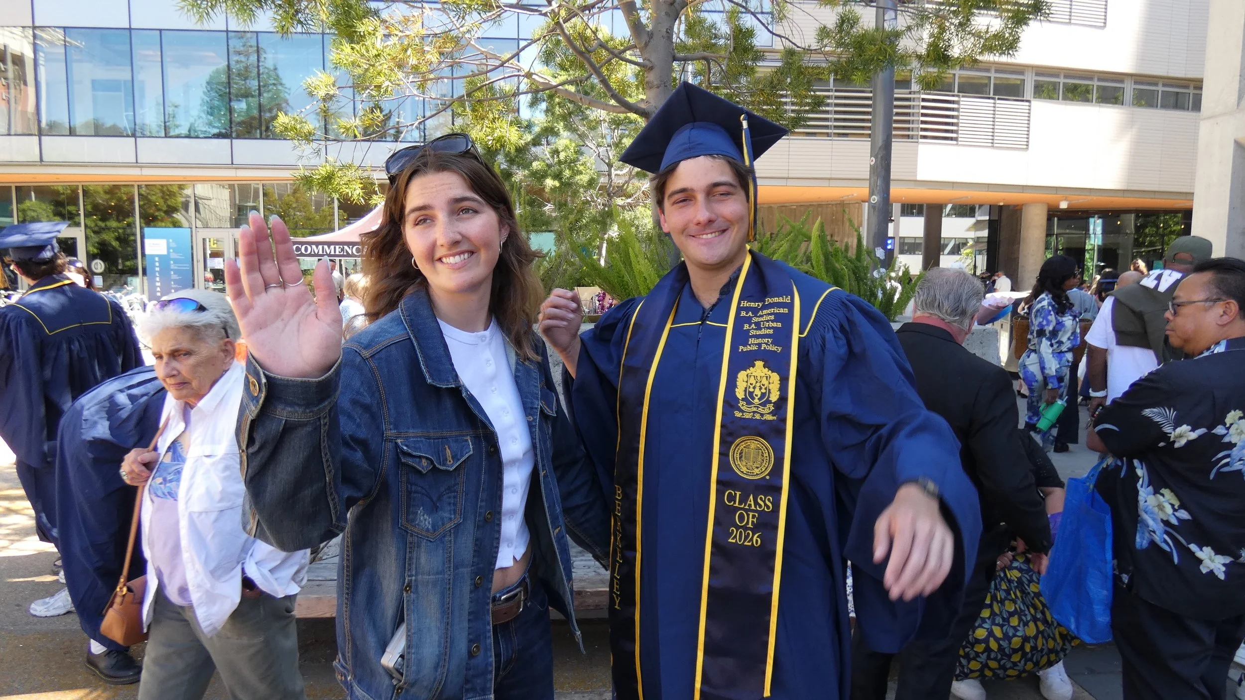 Three young adults celebrating graduation outdoors, with a graduate in a blue cap and gown, smiling and waving, surrounded by other attendees.