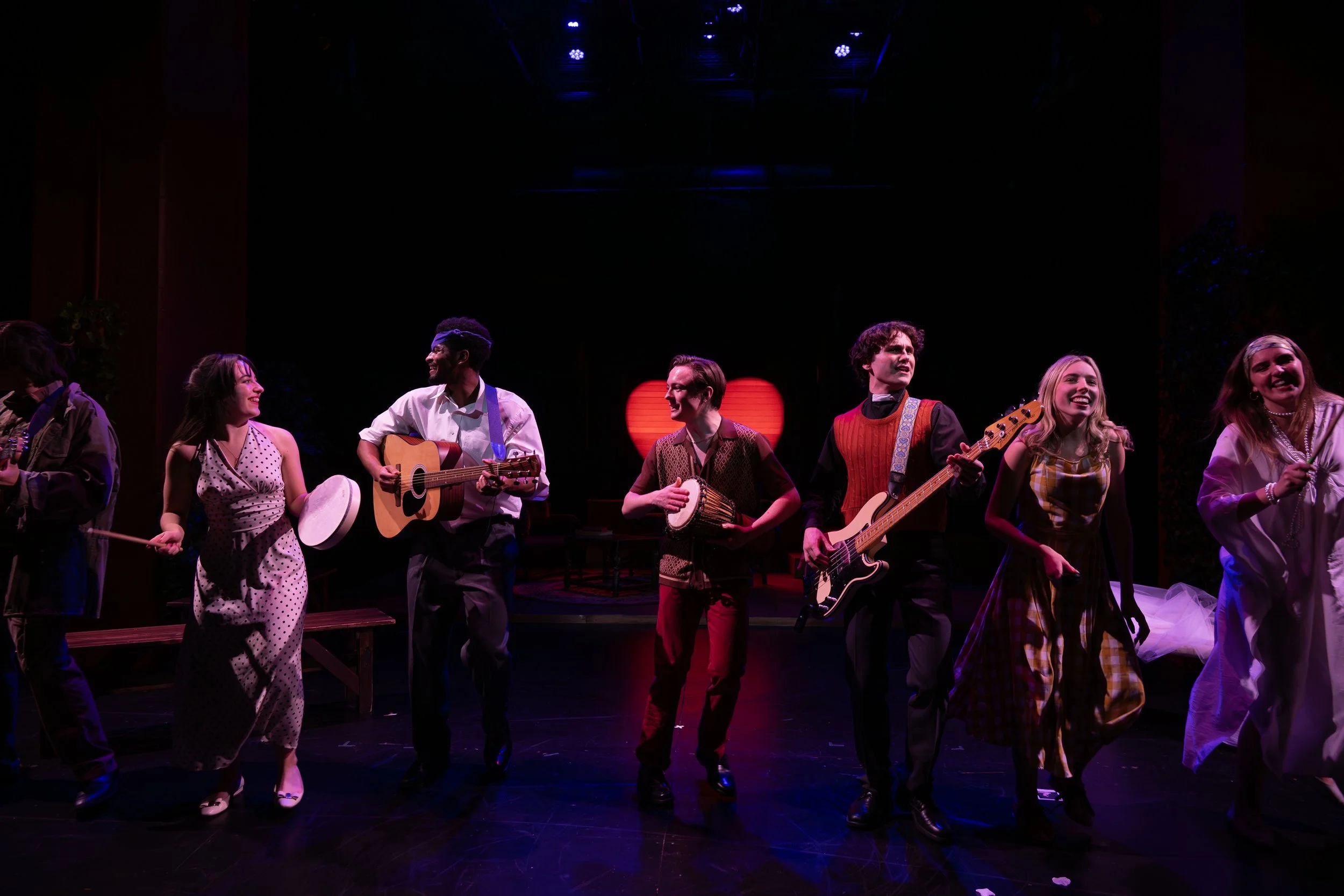 Group of performers on stage with musical instruments, smiling and interacting, with a dark background and red heart-shaped light in the center.