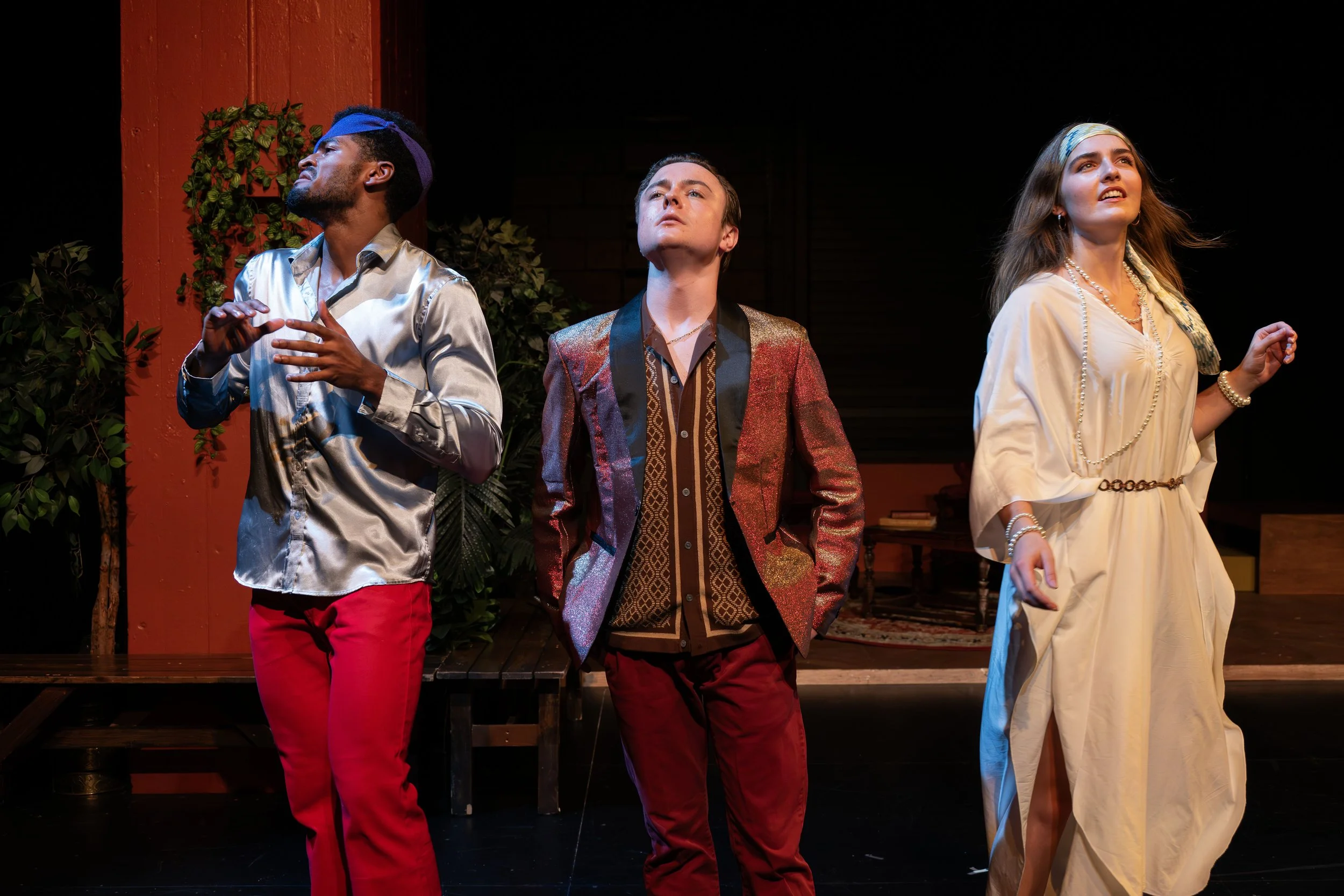 Three actors on stage, one man wearing a shiny silver shirt and red pants, another man in a red and gold blazer, and a woman in a cream-colored dress, performing in a play with dark background and stage setting.
