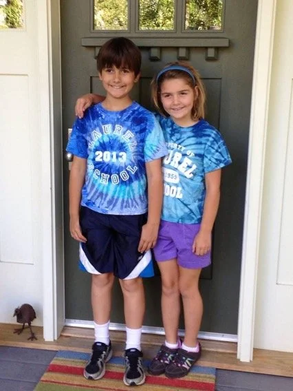 Two children, a boy and a girl, standing together in front of a door indoors, smiling for the camera.