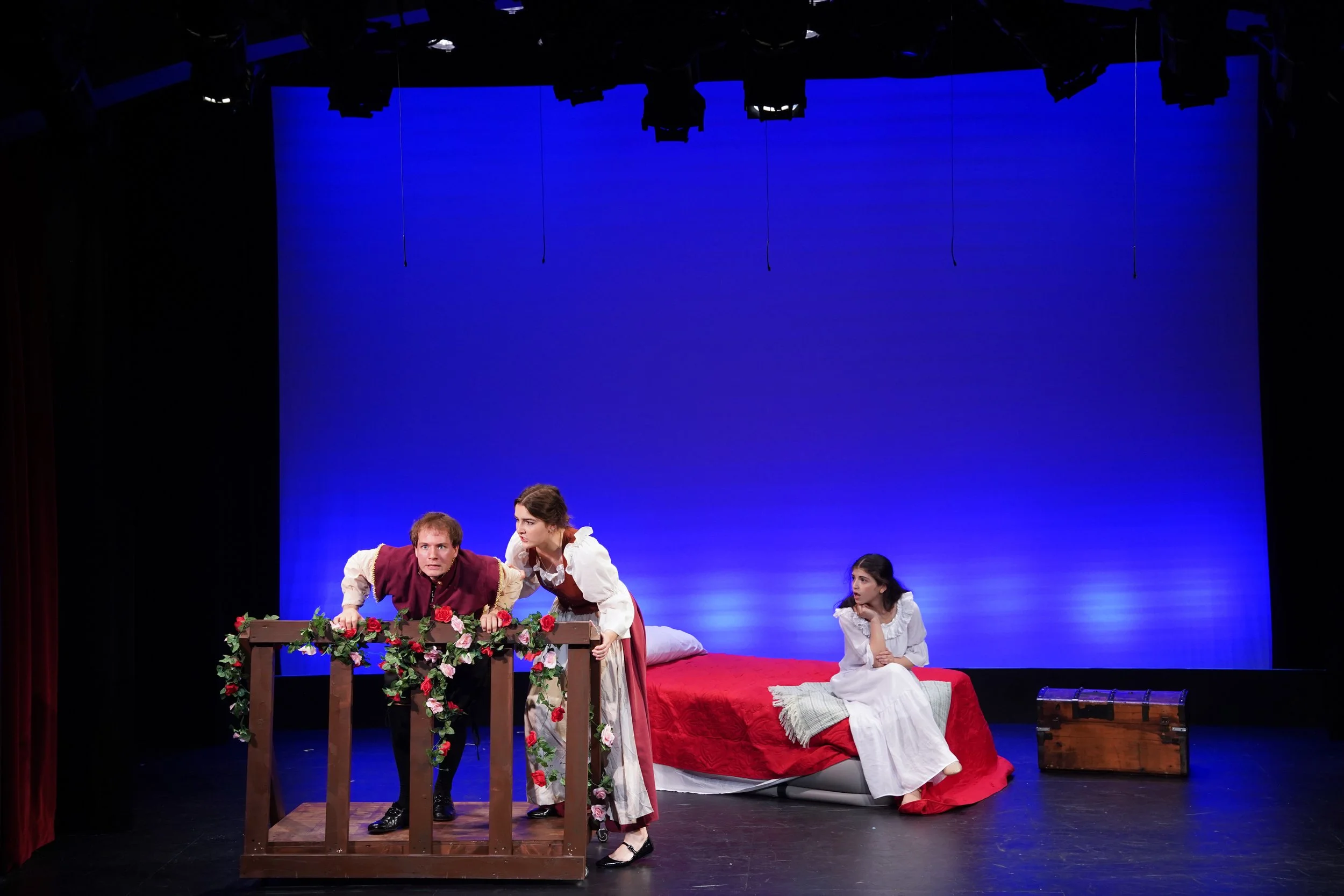 The image shows a scene from a theatrical play with three actors on stage, two women and one man, dressed in period costumes. The stage background is illuminated in blue, and there is a bed and a small wooden table decorated with flowers on stage.
