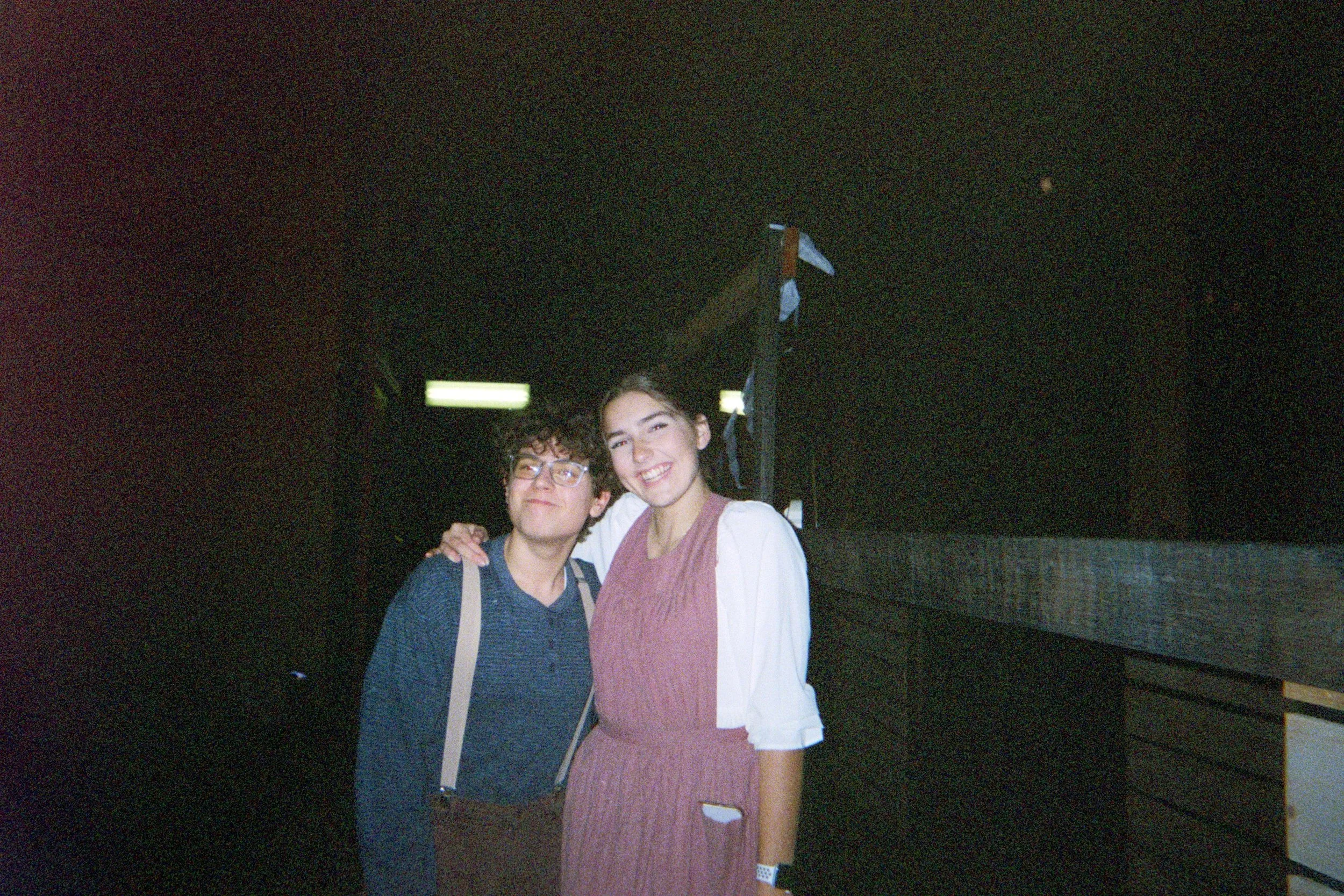 Two young women standing close together, smiling for the camera in an indoor location with dark background and overhead lighting. One woman has curly hair and glasses, while the other has straight hair and is wearing a white and pink dress.