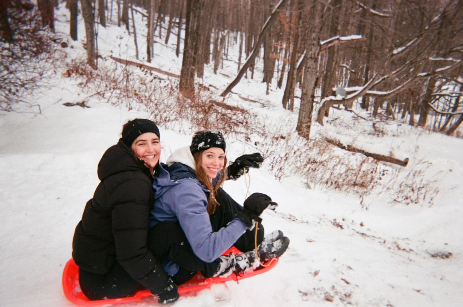 Two young women sitting on a red sled on snow in a wooded area, smiling and enjoying winter.