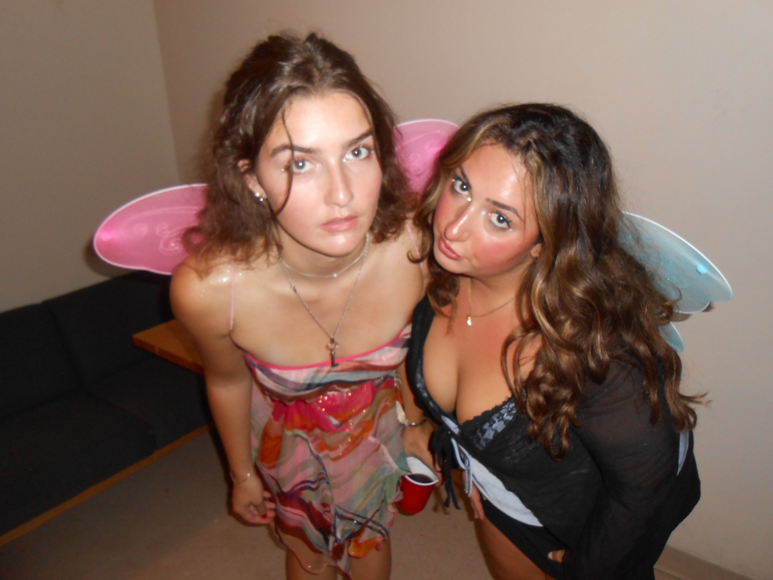 Two women with fairy wings, one with brown curly hair and the other with long wavy hair, posing together indoors.