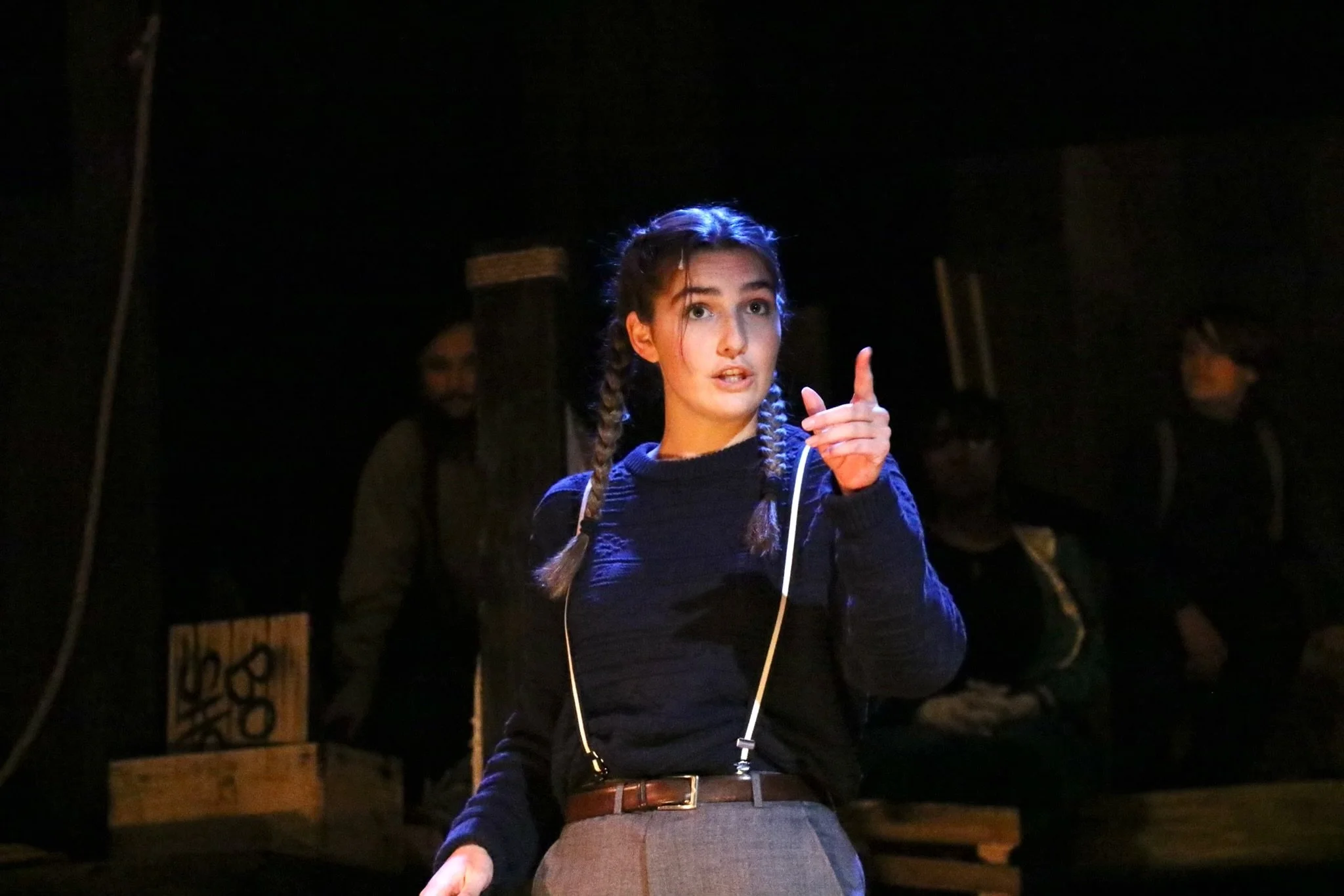 A young woman with braided hair pointing with her right hand while wearing headphones in a dark setting, with two other people in the background.