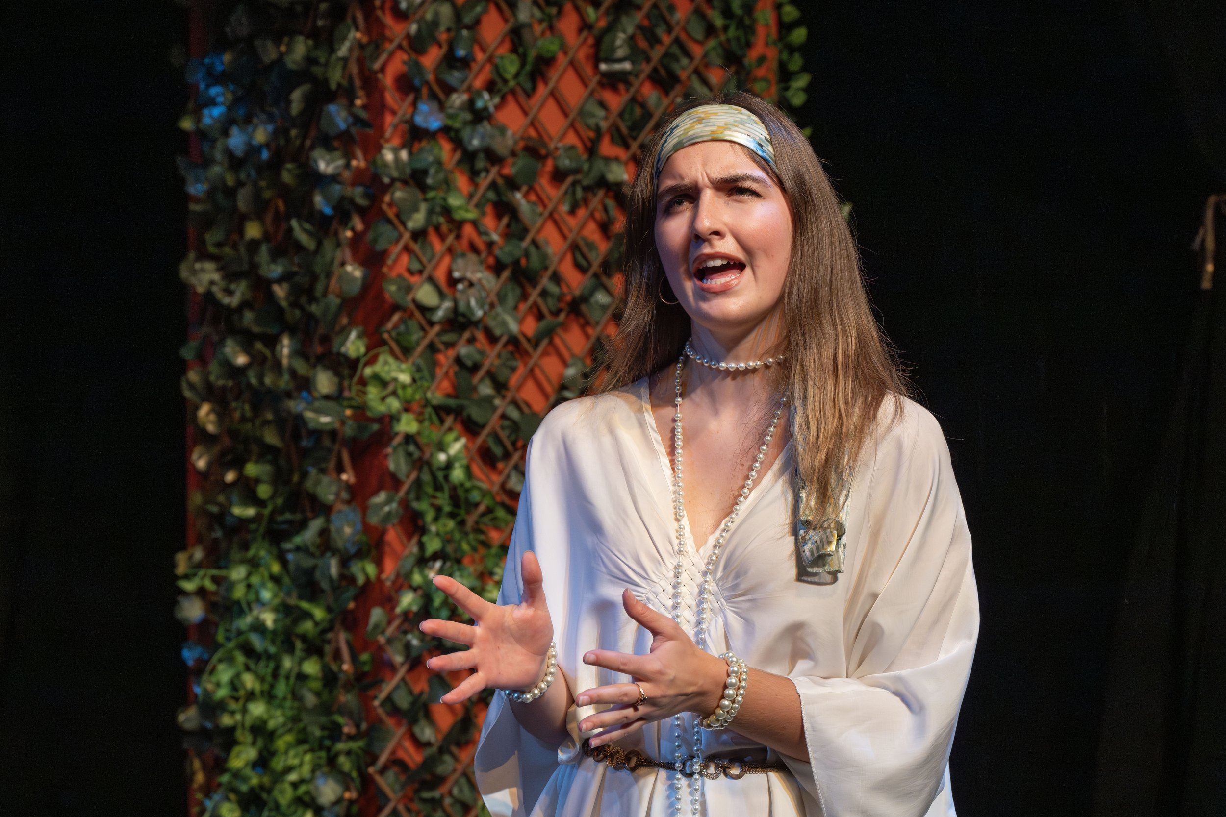 A woman with long brown hair wearing a blue headband, pearl necklace, and bracelet, stands in front of a black background with a red lattice covered in green leaves and plants. She appears to be speaking or singing, with her mouth open and her hands 