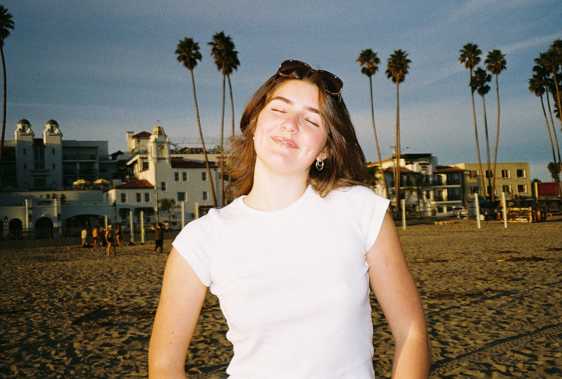 A woman with shoulder-length brown hair, closed eyes, and a slight smile, wearing a white t-shirt, standing on a beach during sunset with palm trees and buildings in the background.