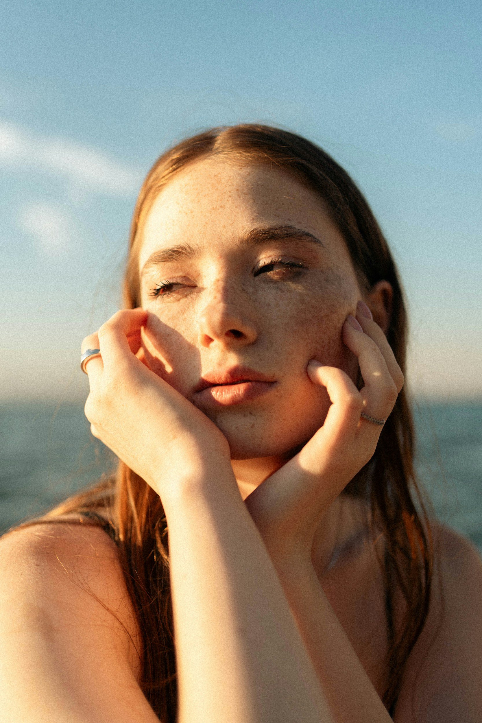 Close-up of a woman with red hair and freckles, eyes closed, squinting in sunlight, touching her face with hands on face and cheeks, outdoors with blue sky