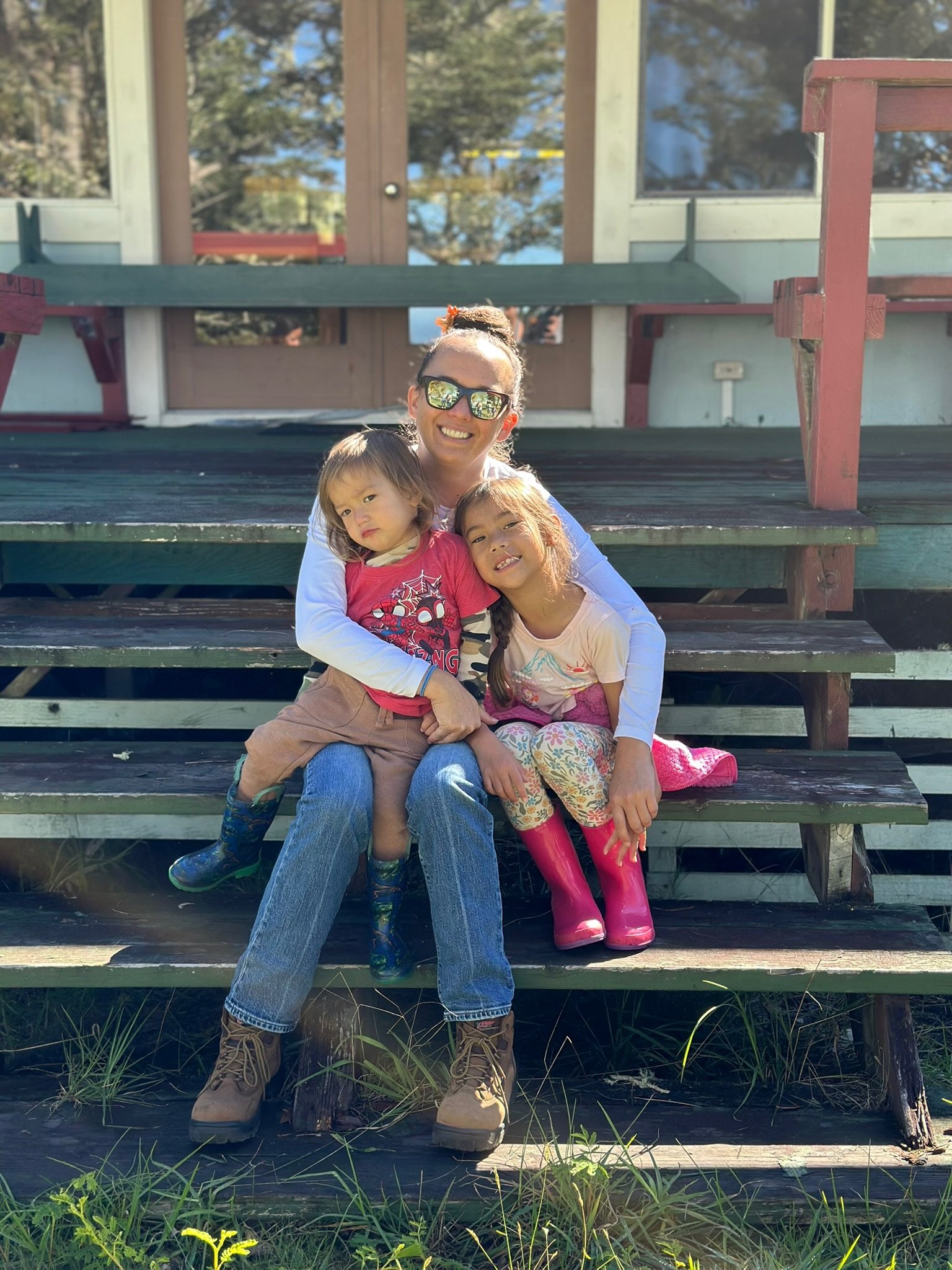 A woman with sunglasses smiling and two young girls sitting on wooden steps outdoors. One girl looks serious, the other is smiling. The setting appears to be a porch or deck of a house with trees and blue sky reflected in the background.