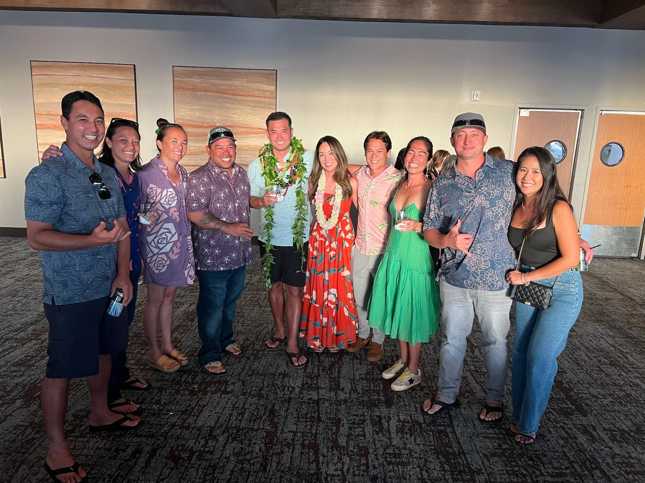 A group of eleven people in colorful, casual, summer clothing standing together indoors, some holding drinks, with happy expressions, posing for a photo at a social gathering.