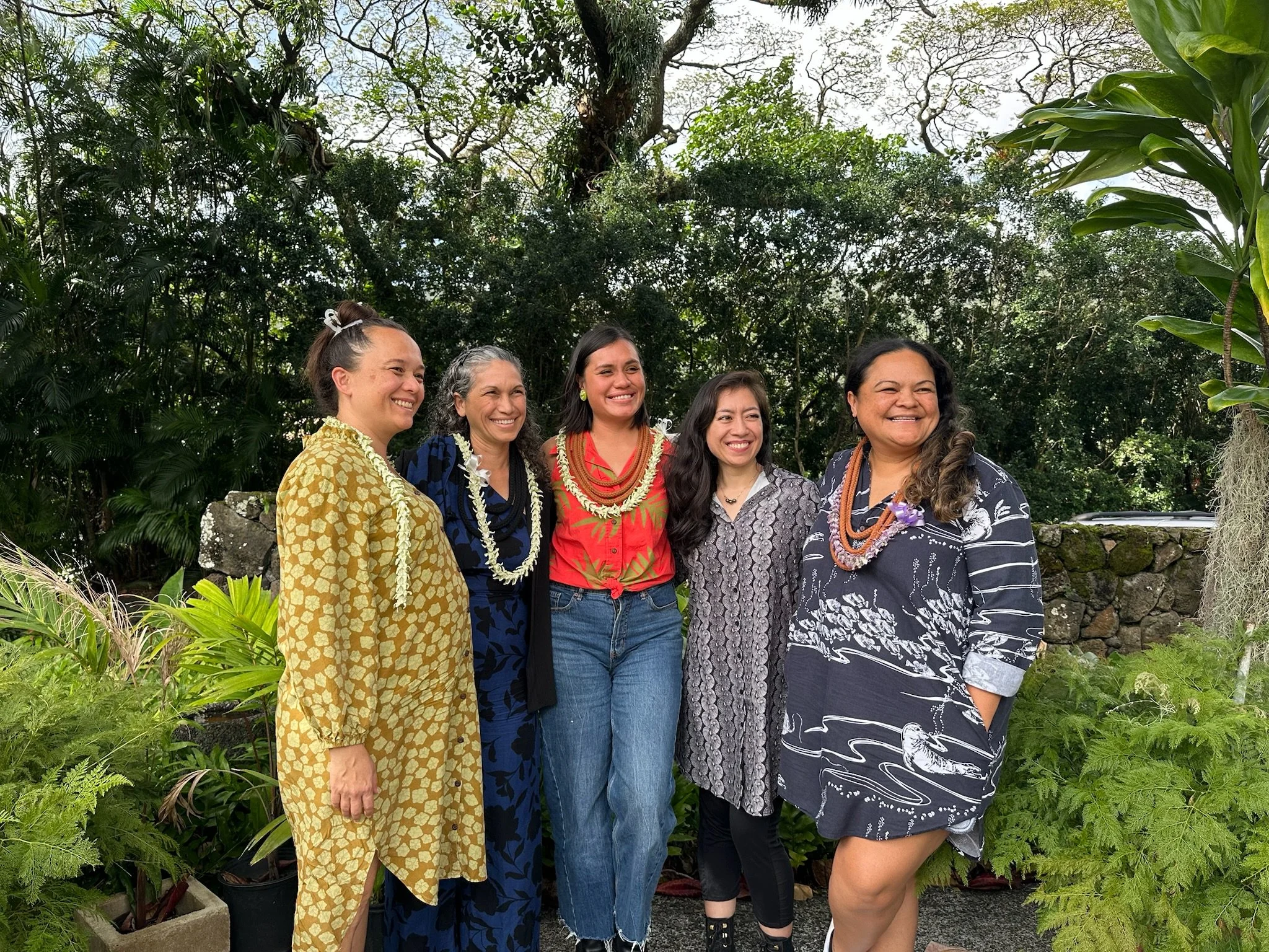 Five women in colorful clothing and leis standing together outdoors in a lush garden.