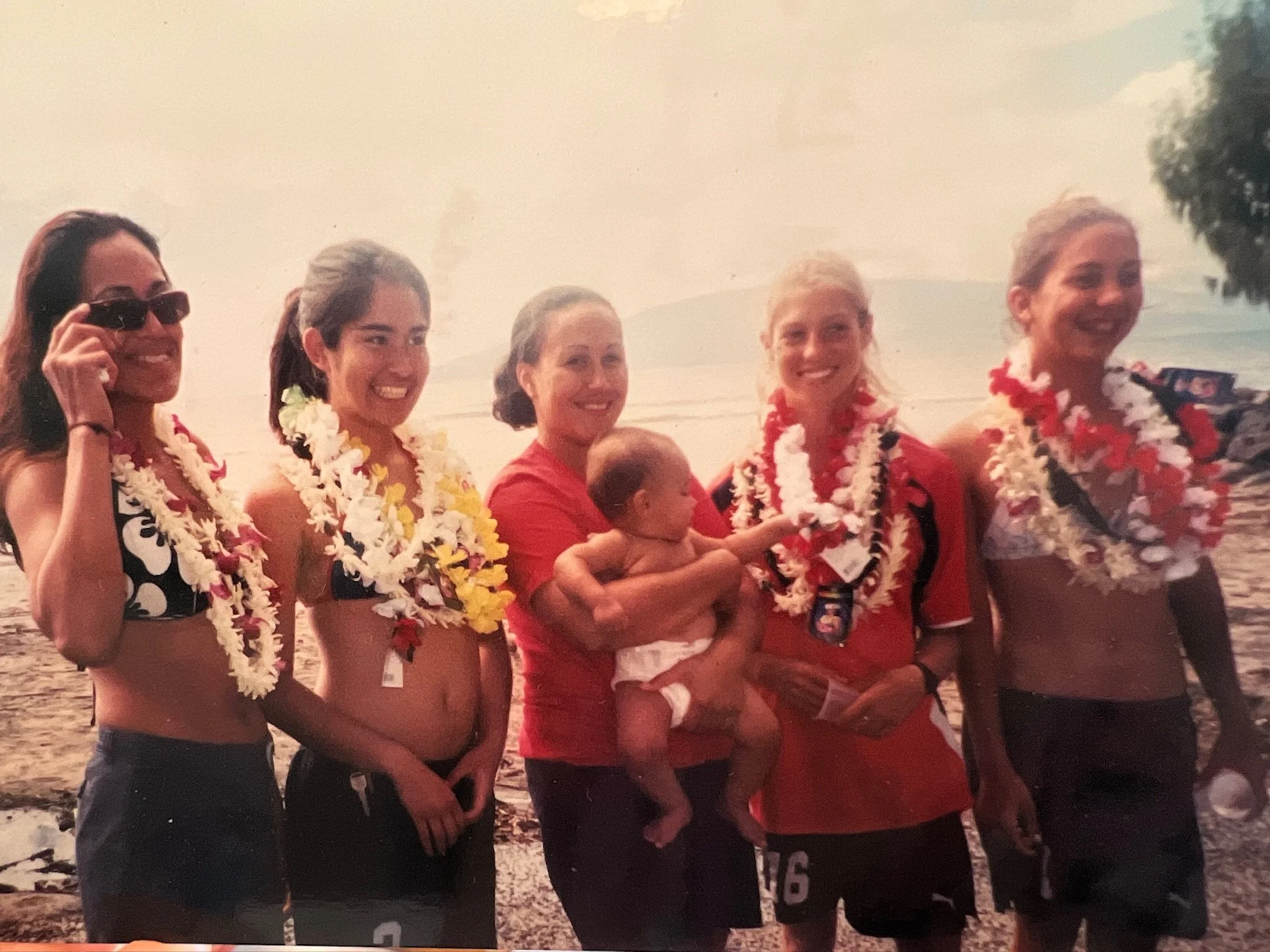 A group of five women and a child on a beach during sunset, wearing leis and casual summer clothes, smiling and posing for the photo.
