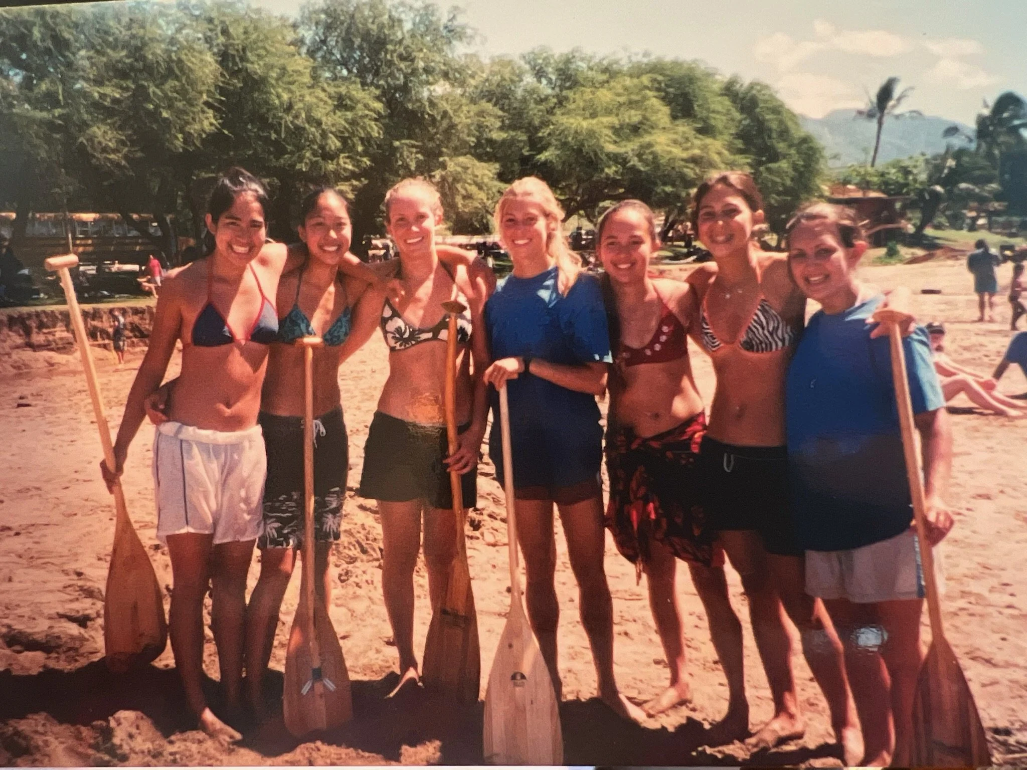 Group of seven women in swimsuits standing outdoors on sandy ground, smiling, some holding paddles, with lush green trees and a clear sky in the background.