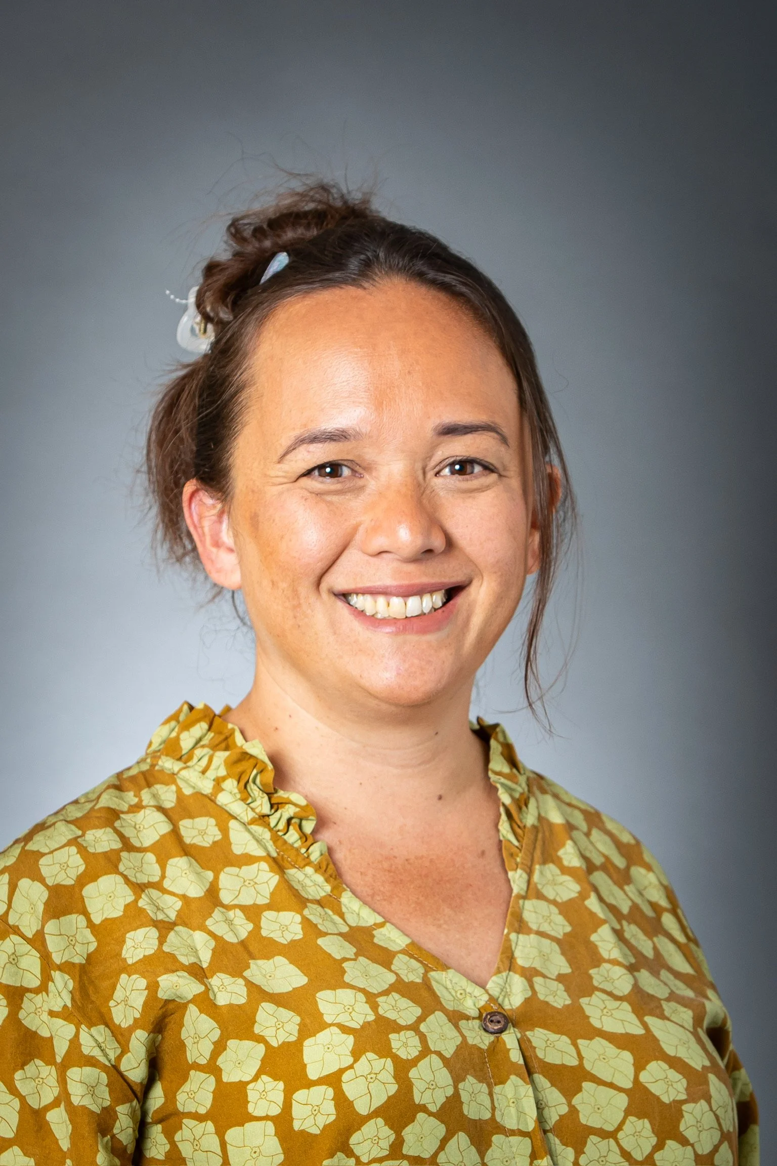 A woman with brown hair styled in an updo, smiling, wearing a mustard yellow blouse with a cream-colored floral pattern, against a neutral gray background.