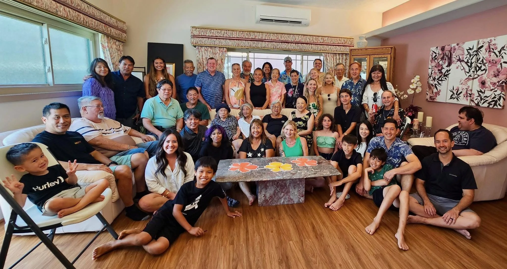 A large diverse group of people of various ages gathered in a cozy living room for a group photo, sitting on sofas and standing behind, smiling and posing for the camera.