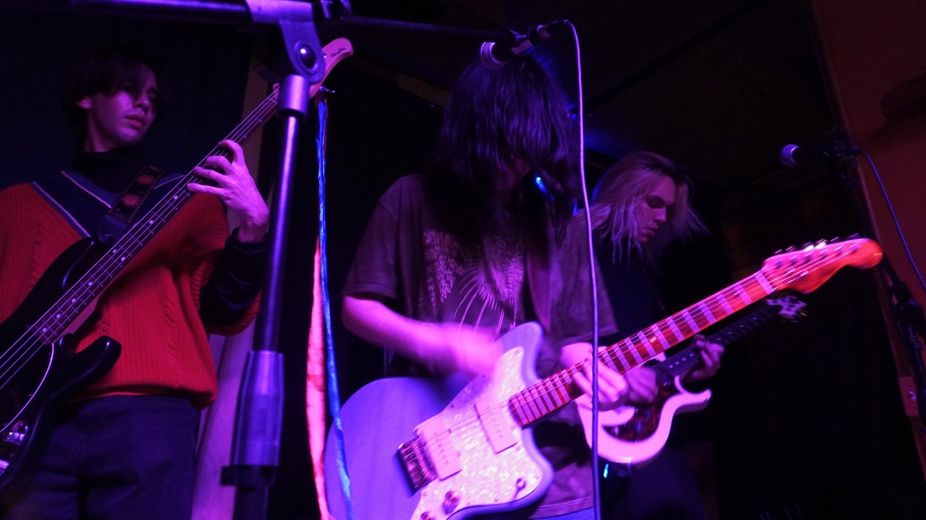 Three musicians playing guitars on stage with purple and pink lighting, microphones, and a black background.