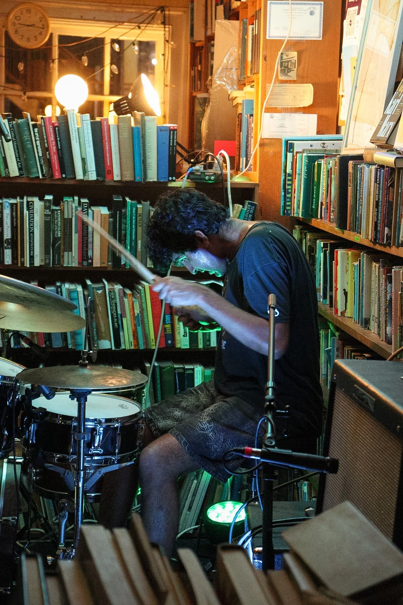A person playing drums in a room filled with bookshelves, with warm lighting and visible paperwork on the walls.