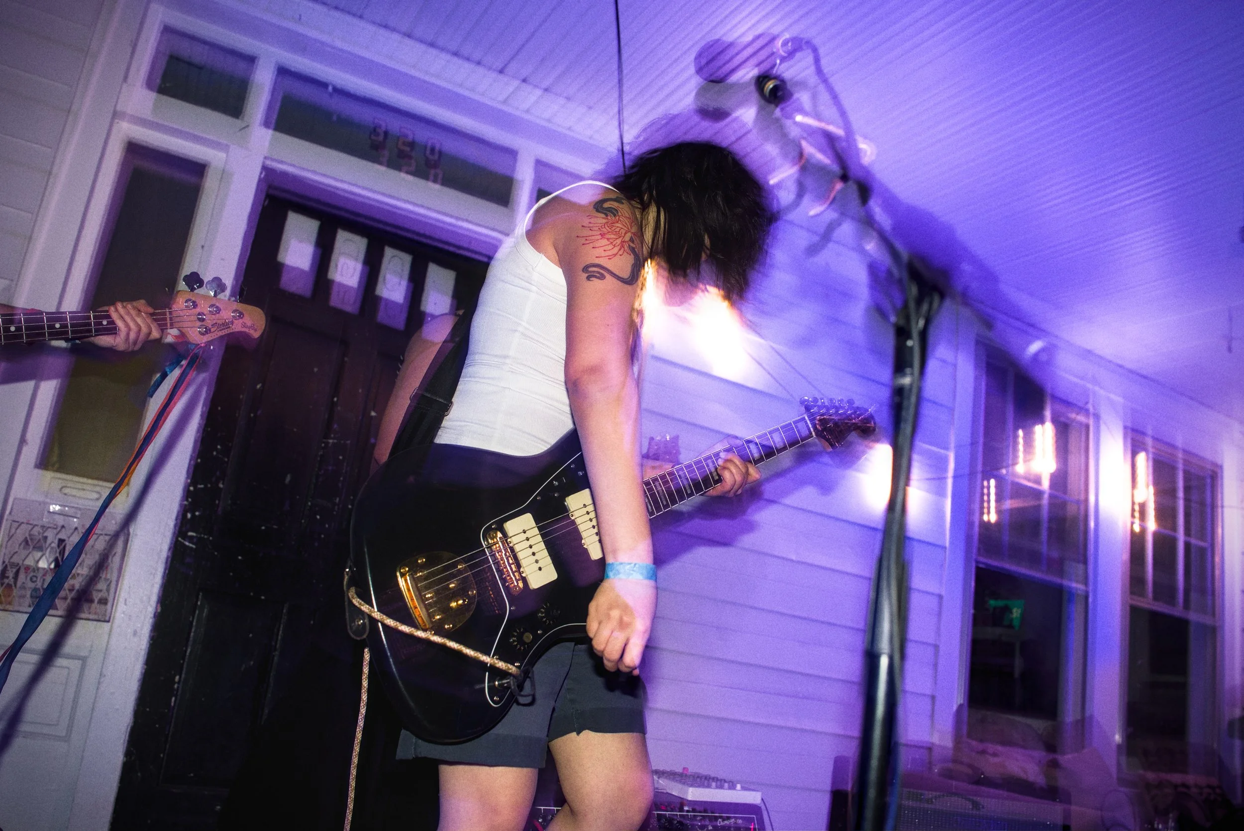 A woman with dark hair playing an electric guitar during a live performance at night, illuminated by blue and purple lighting, with other band members partially visible in the background.