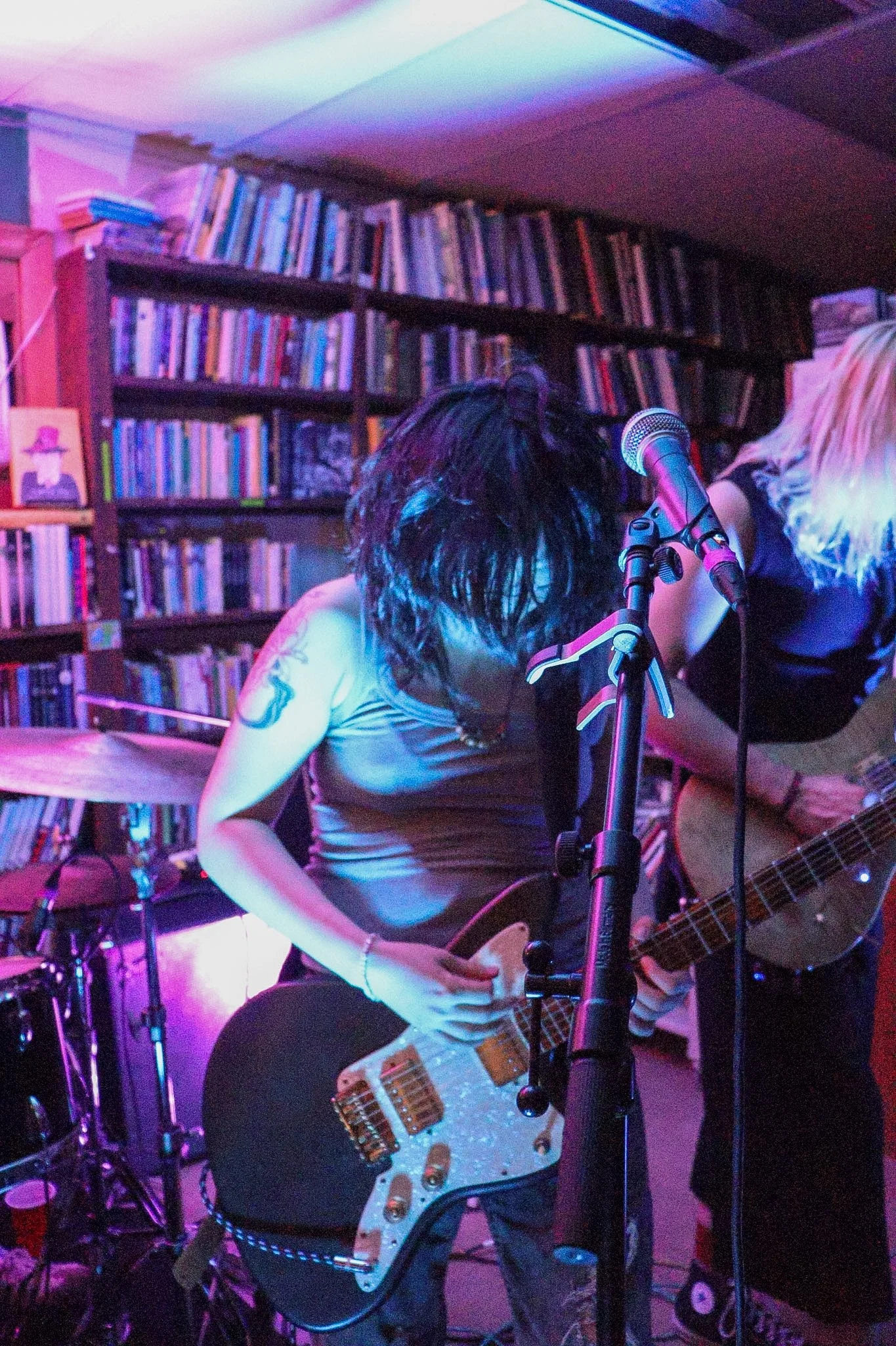 Musician playing an electric guitar in a room with bookshelf background, colorful lighting, and a microphone.