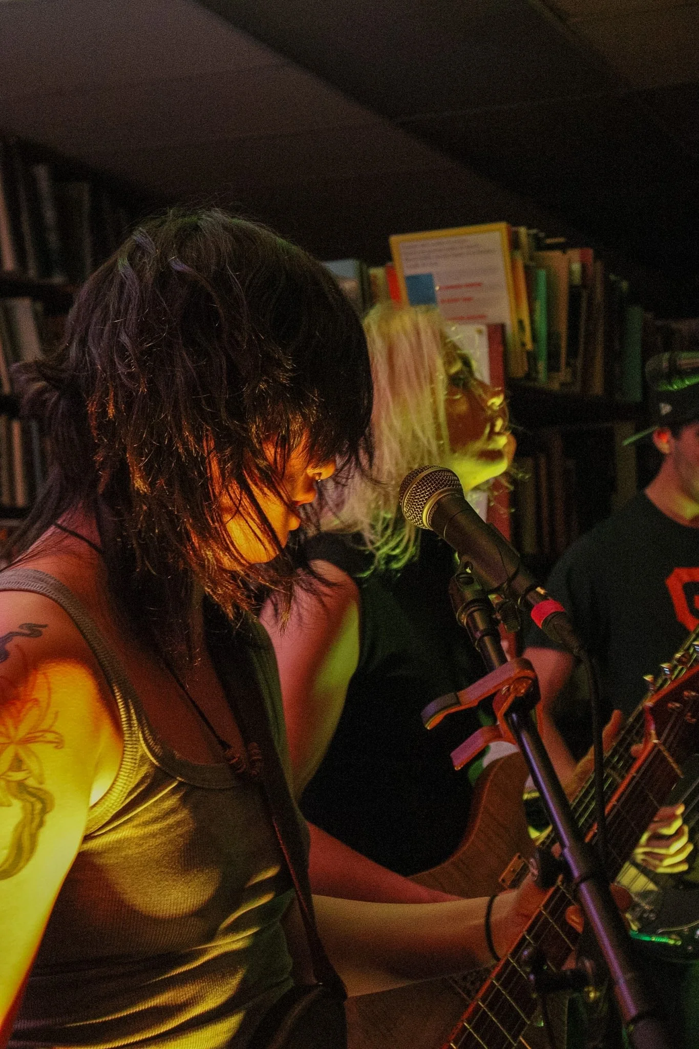 A woman with black hair singing into a microphone while playing an acoustic guitar in a dimly lit room with bookshelves in the background.