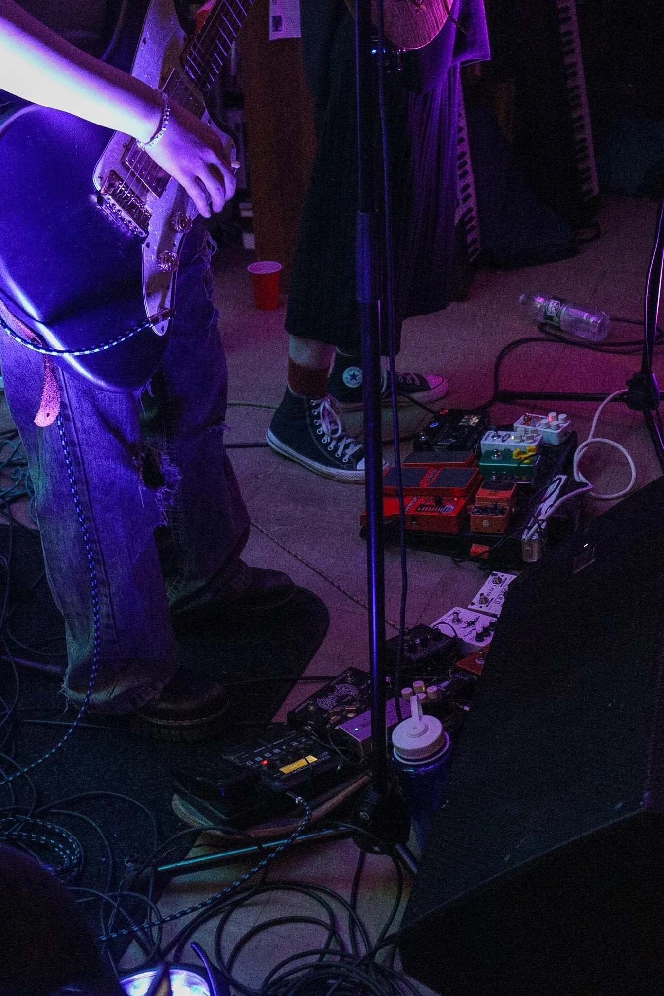 A person playing an electric guitar on a dimly lit stage with purple lighting. The person is wearing distressed jeans, a bracelet, and a ring. Various music equipment, pedals, and cables are scattered on the floor. A red cup and a water bottle are al