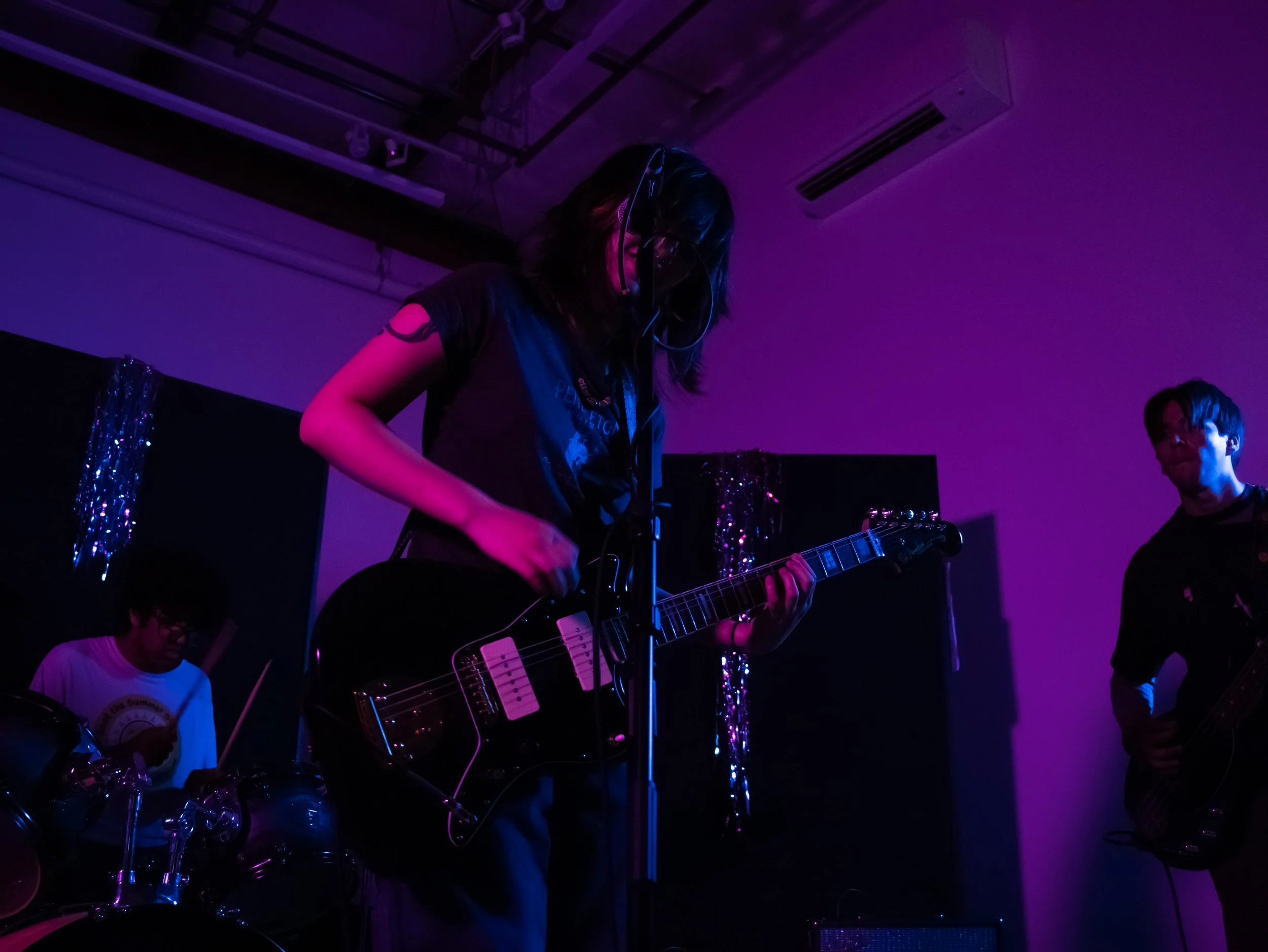 A female musician wearing headphones playing an electric guitar on stage in a dimly lit venue with purple lighting and shiny decorations in the background.