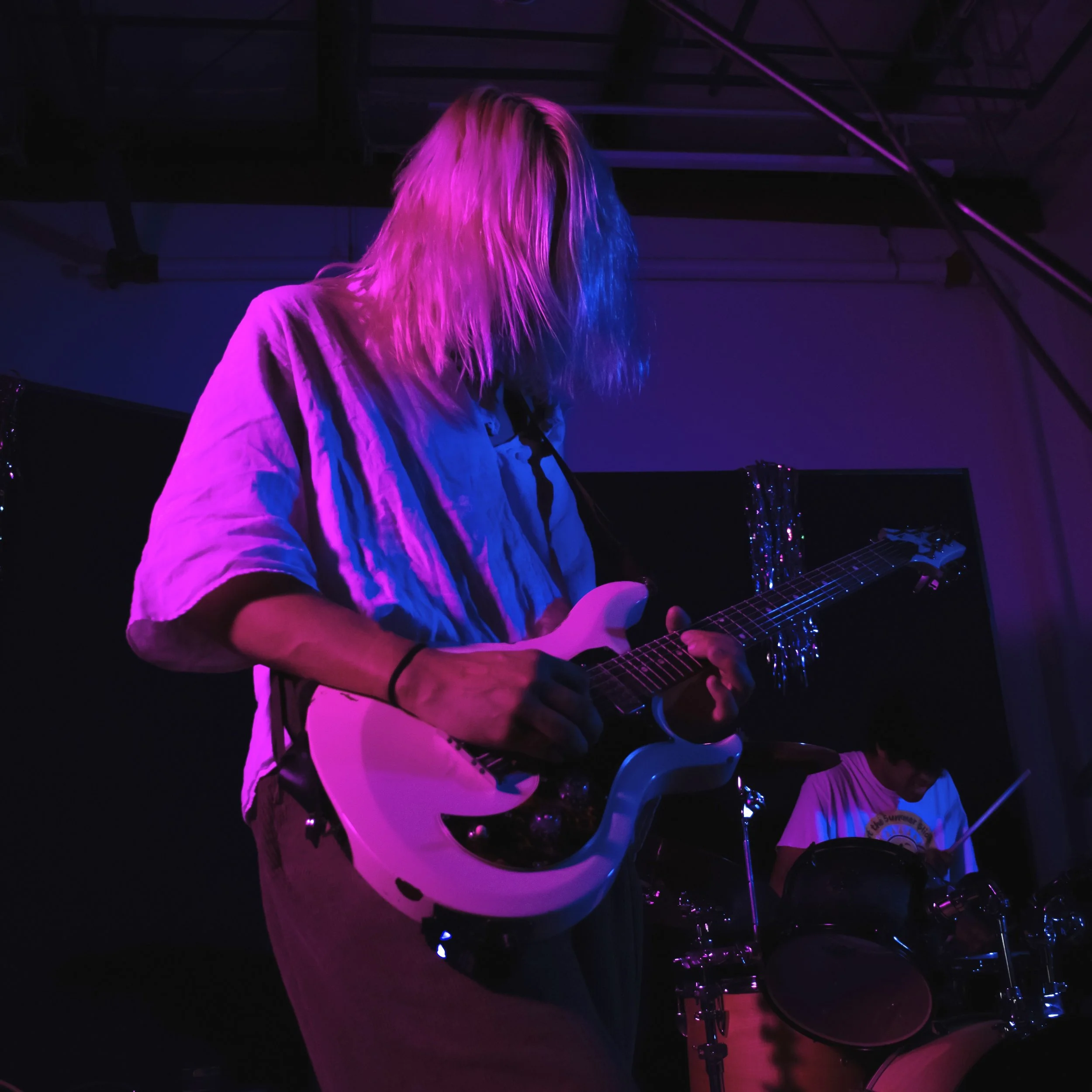 A person with long blonde hair, wearing a white jacket, playing an electric guitar under purple and pink stage lights.