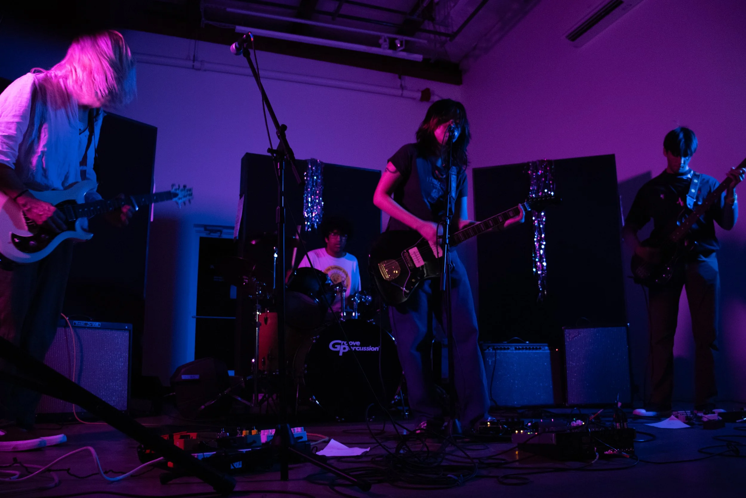 Band performing on stage with purple and blue lighting, three guitarists and a drummer visible, sound equipment on stage.