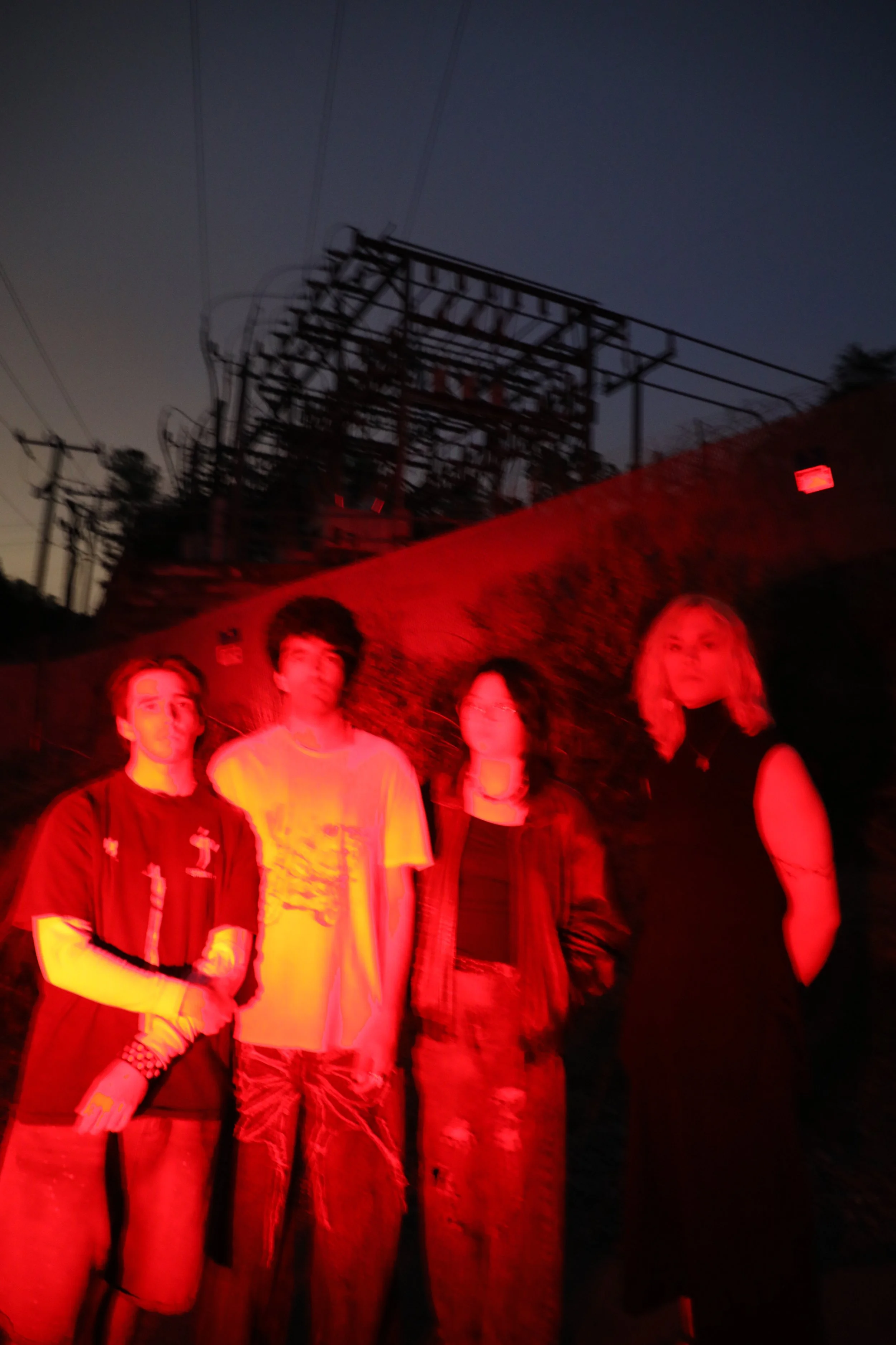 Five people standing outdoors at night, illuminated by red light, with power lines and a metal structure in the background.