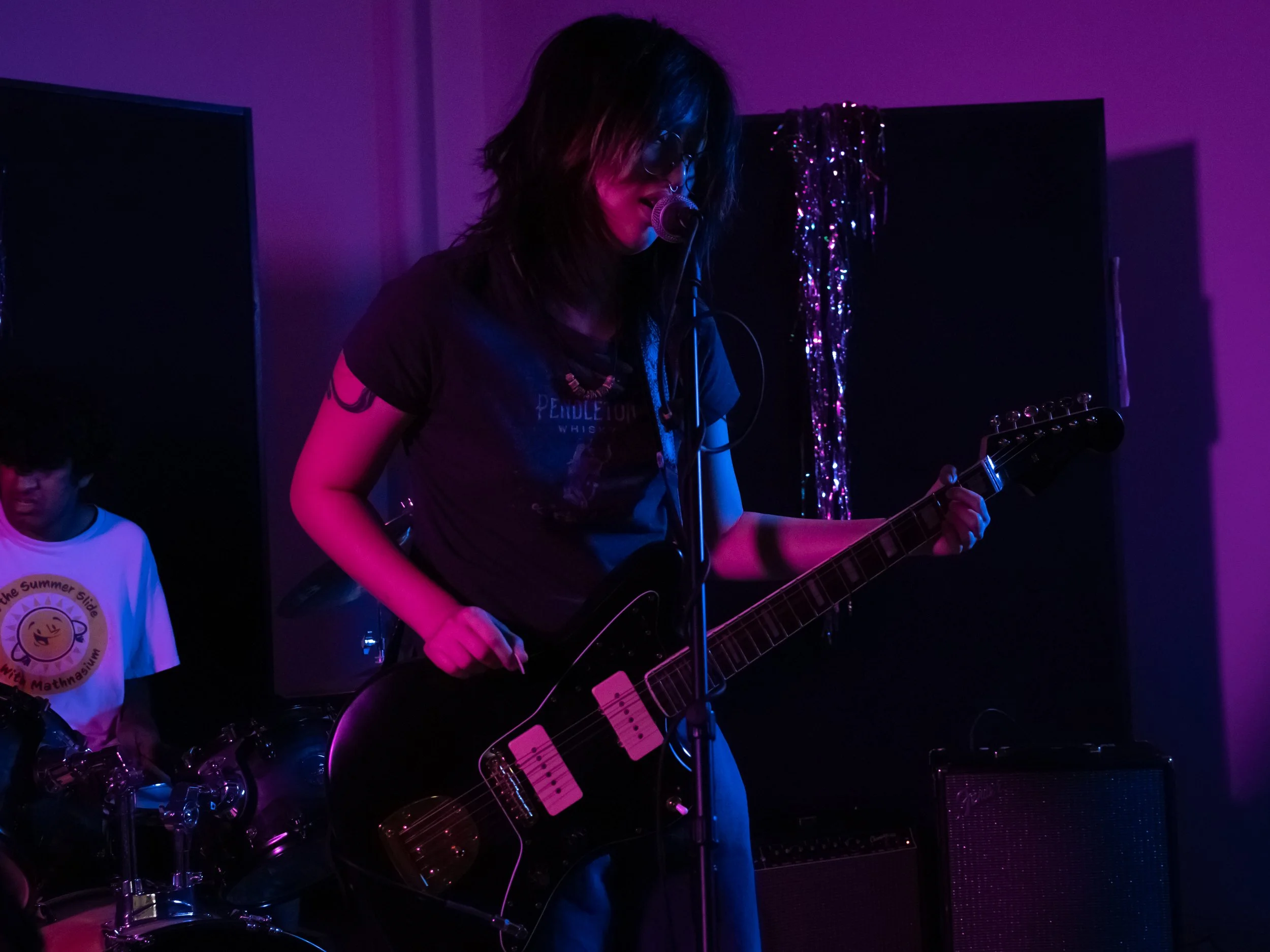 A woman playing an electric guitar and singing into a microphone during a performance, with colored stage lighting.