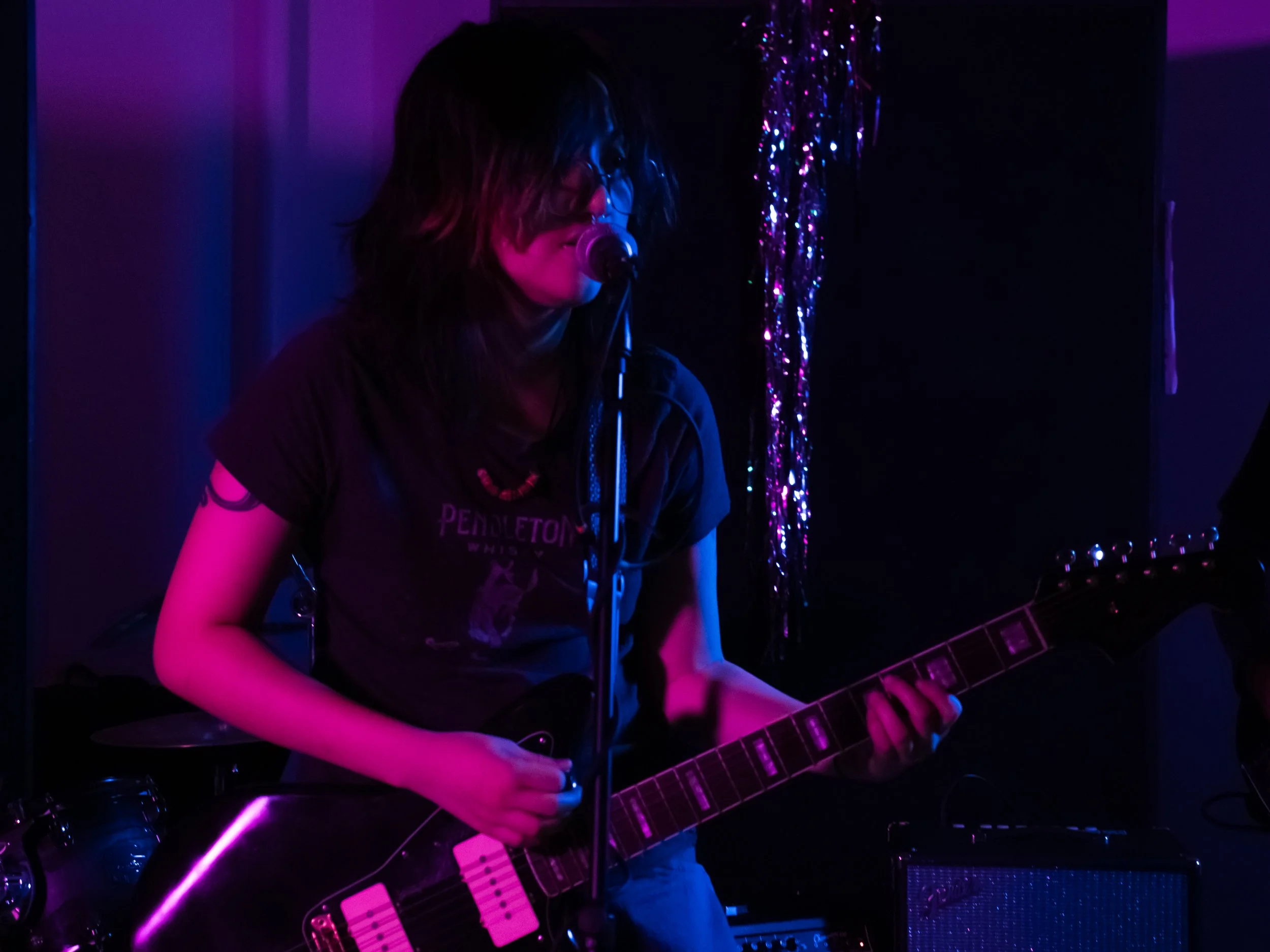 A person with dark hair and glasses singing into a microphone while playing an electric guitar in a dark room illuminated by pink and purple stage lights.