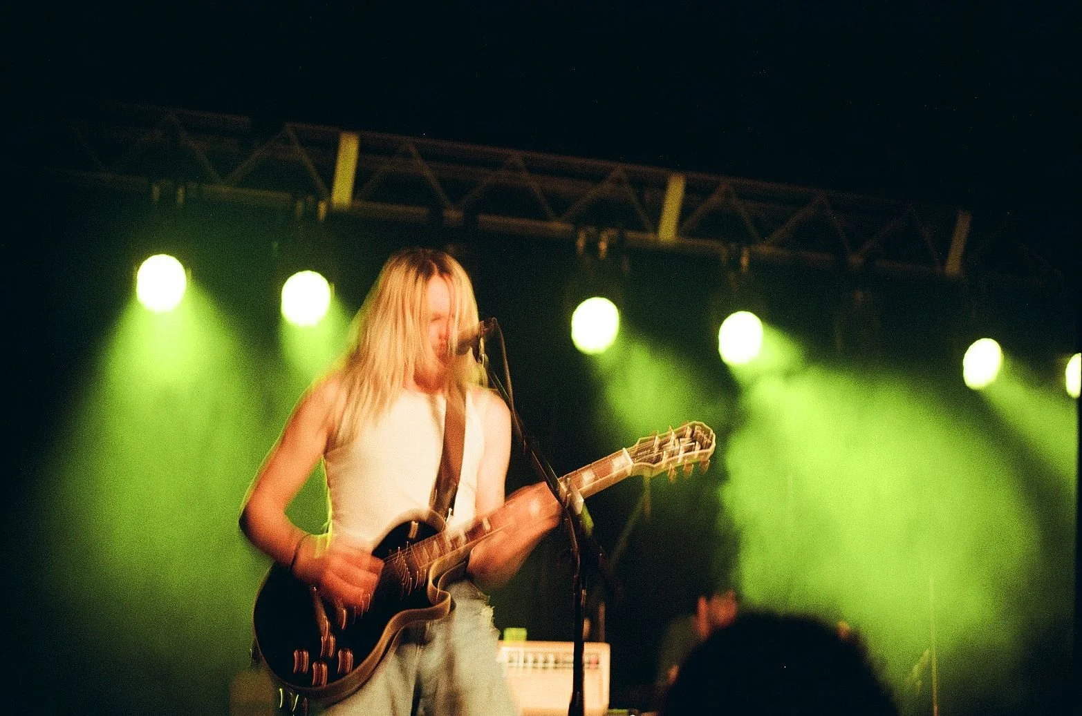 A female musician with long blonde hair performing on stage, playing an electric guitar and singing into a microphone, illuminated by green stage lights at night.