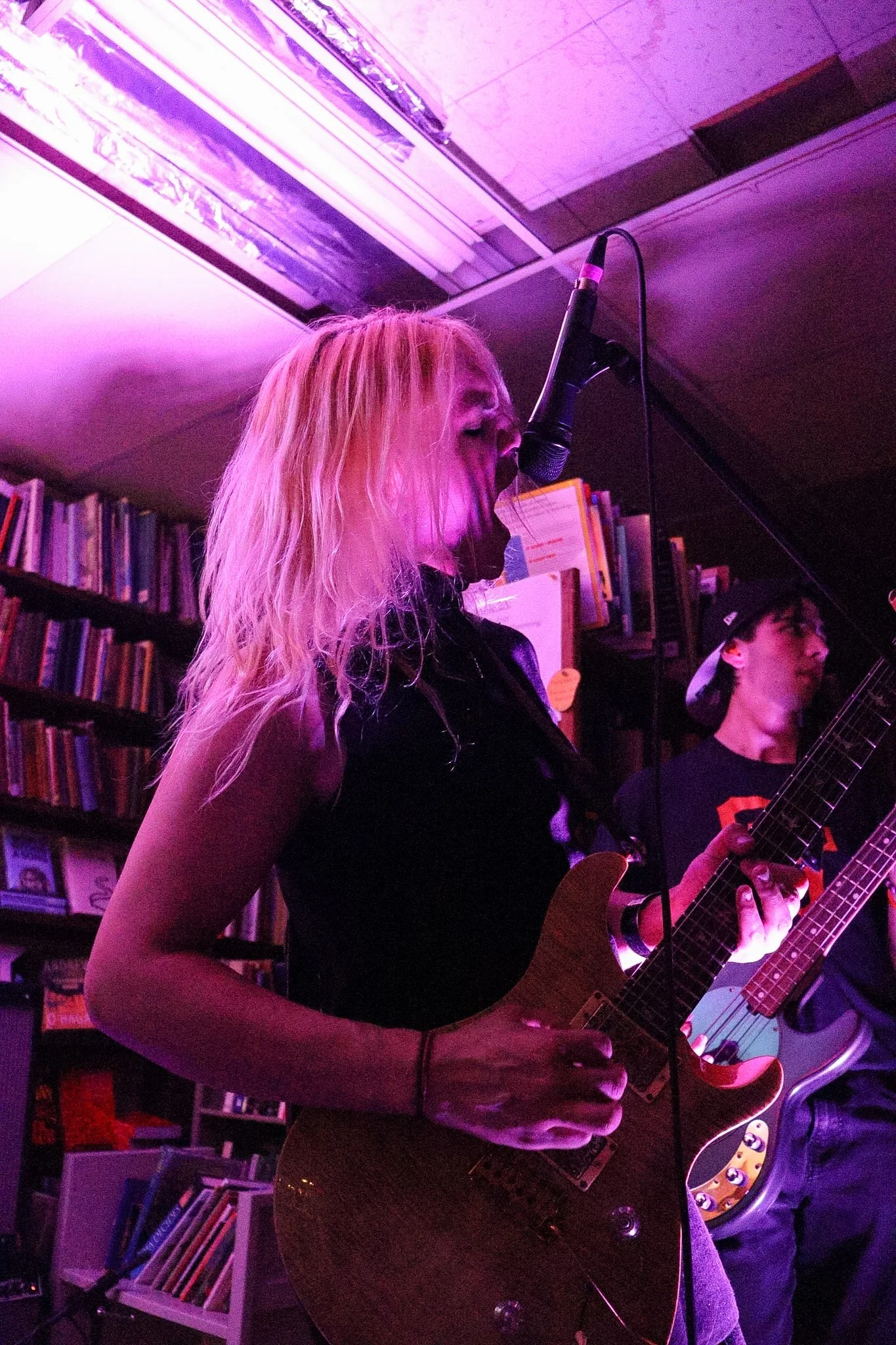 A woman with long blond hair singing into a microphone and playing an electric guitar in a room with purple lighting. Bookshelves are visible in the background.