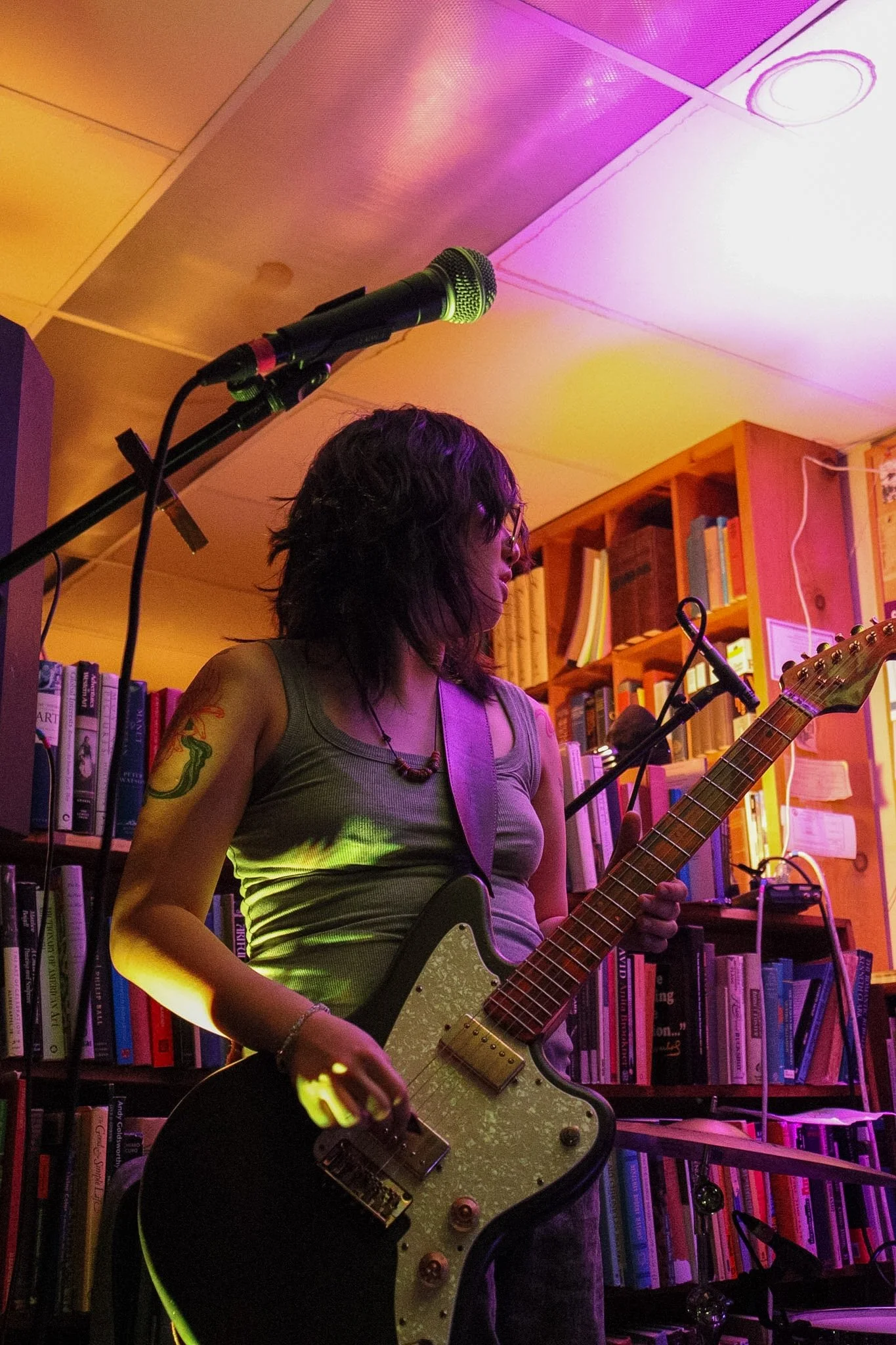 A woman with glasses and tattoos on her arm playing an electric guitar in a cozy room with bookshelves and colorful lighting.