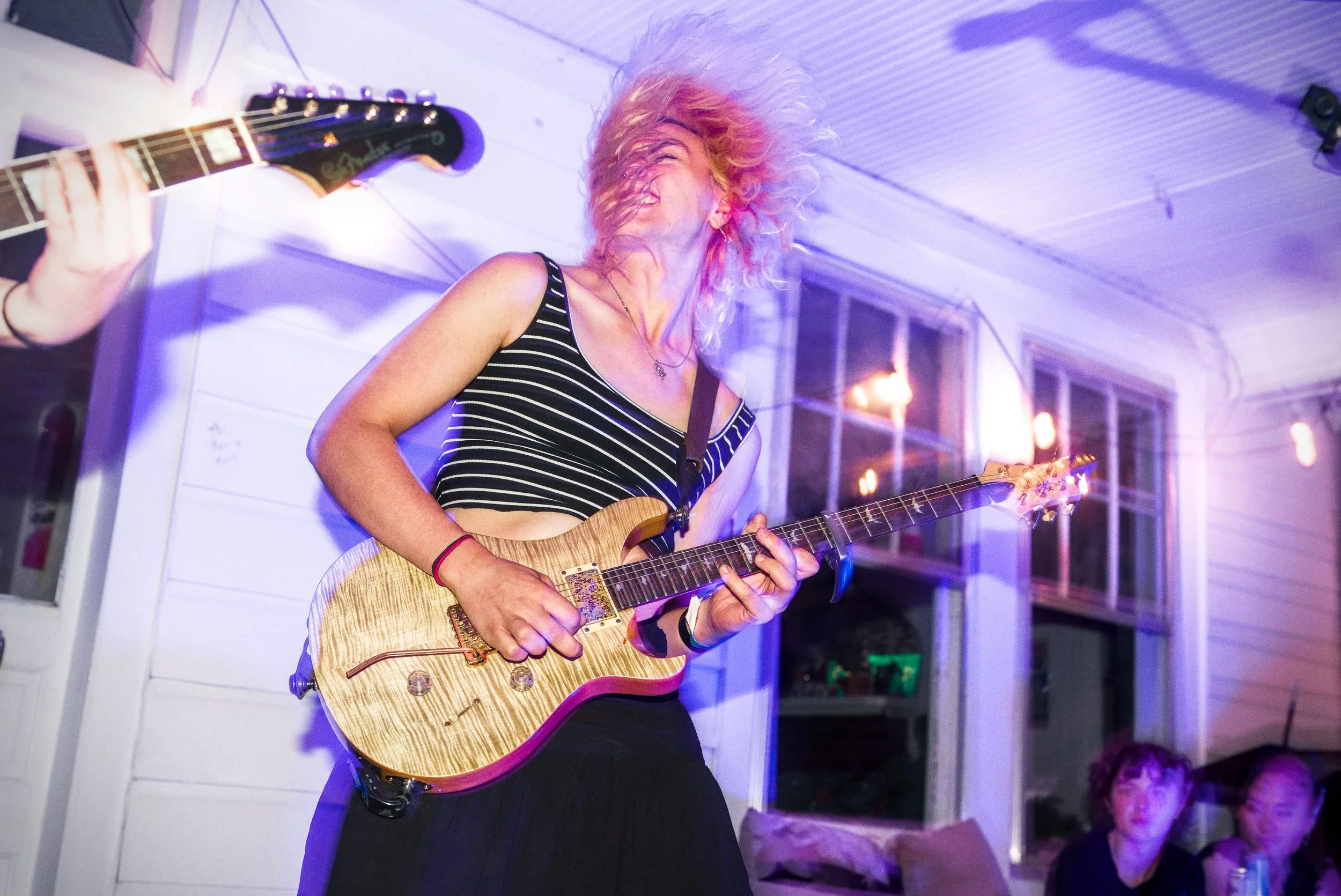 A woman with curly blonde hair is playing an electric guitar at a lively indoor event, smiling and enjoying the music. She is wearing a black and white striped tank top, and there are people in the background watching her perform.